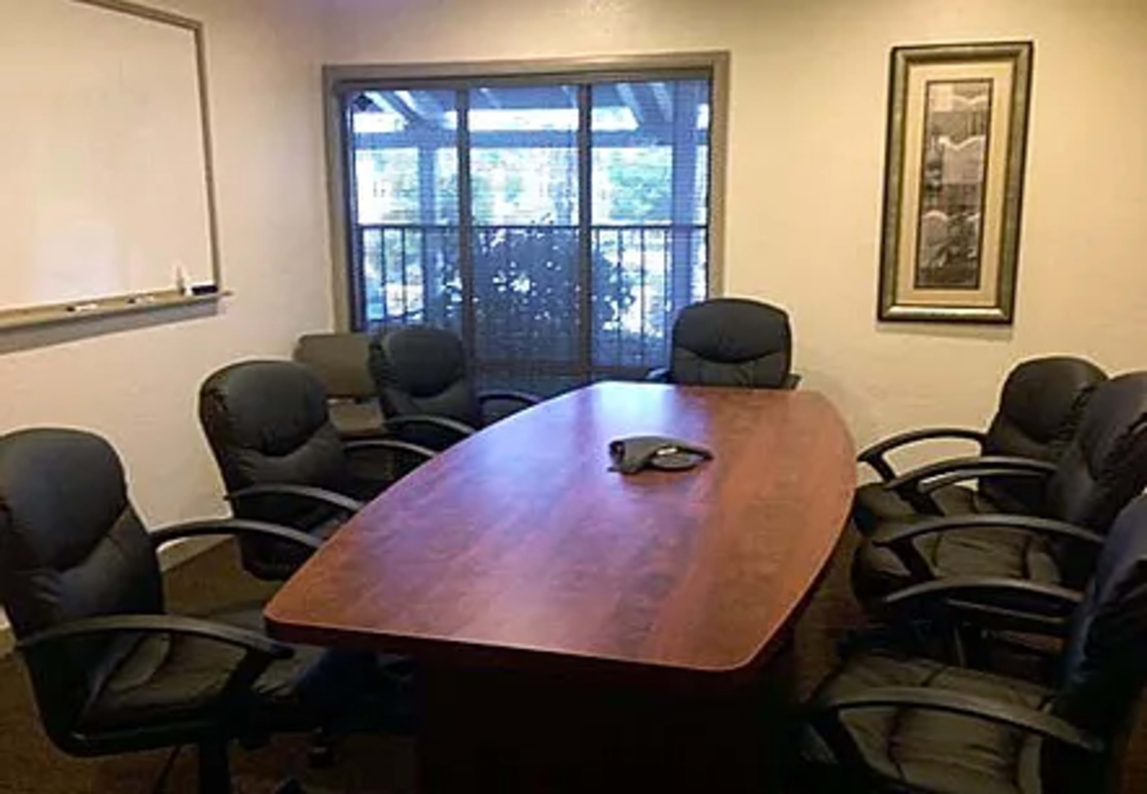 A conference room with a large wooden table surrounded by eight black chairs, and a window displaying greenery outside.
