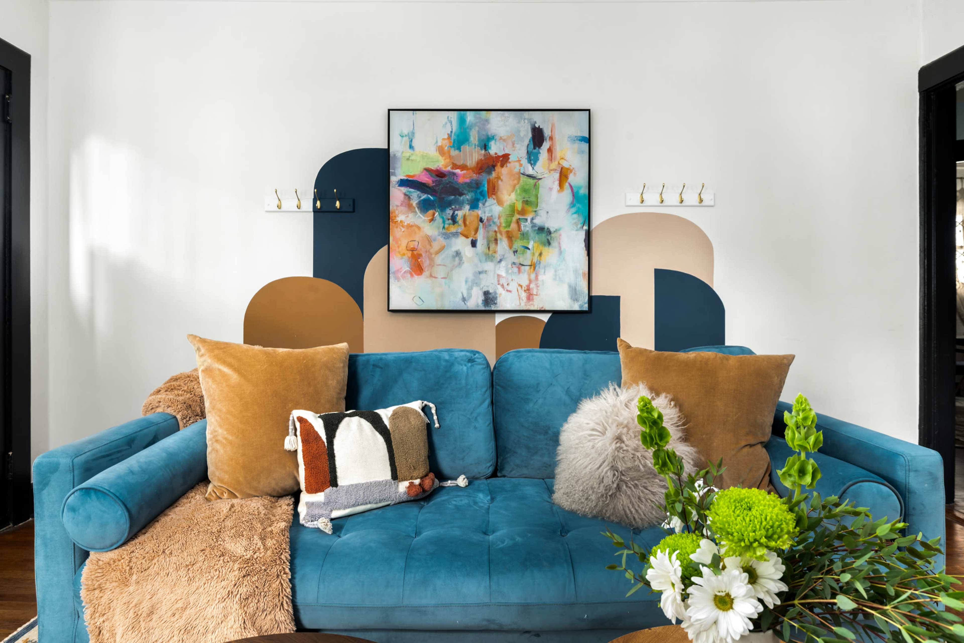 The image shows a stylish living room featuring a blue couch adorned with various decorative pillows, a colorful abstract painting on the wall, and a small table with a floral arrangement.
