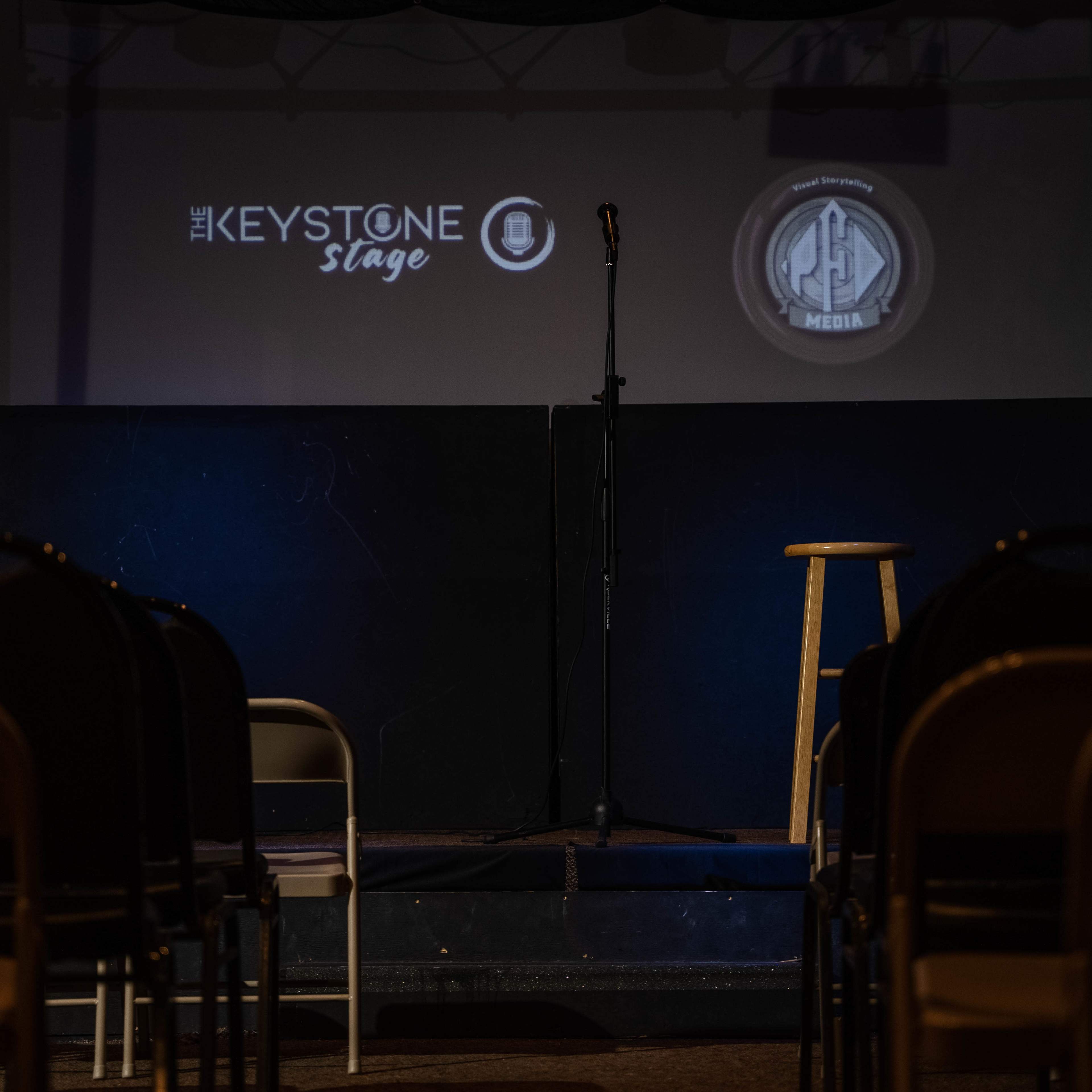 A stage set up for a performance, featuring a microphone on a stand, a wooden stool, and empty chairs arranged in front.