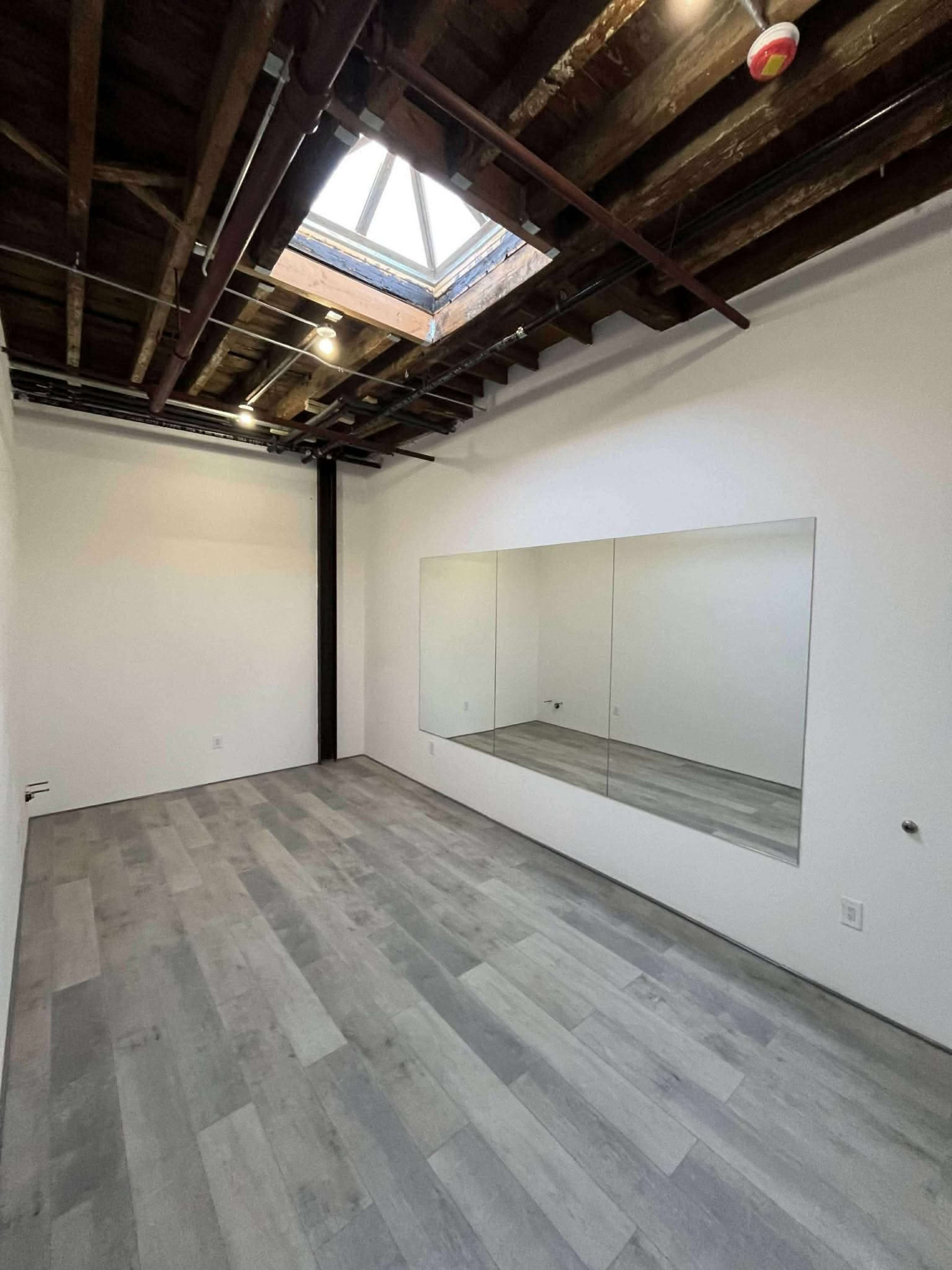 The image shows a spacious room with wooden floors, exposed beams, and large mirrors on one wall, featuring a skylight above.