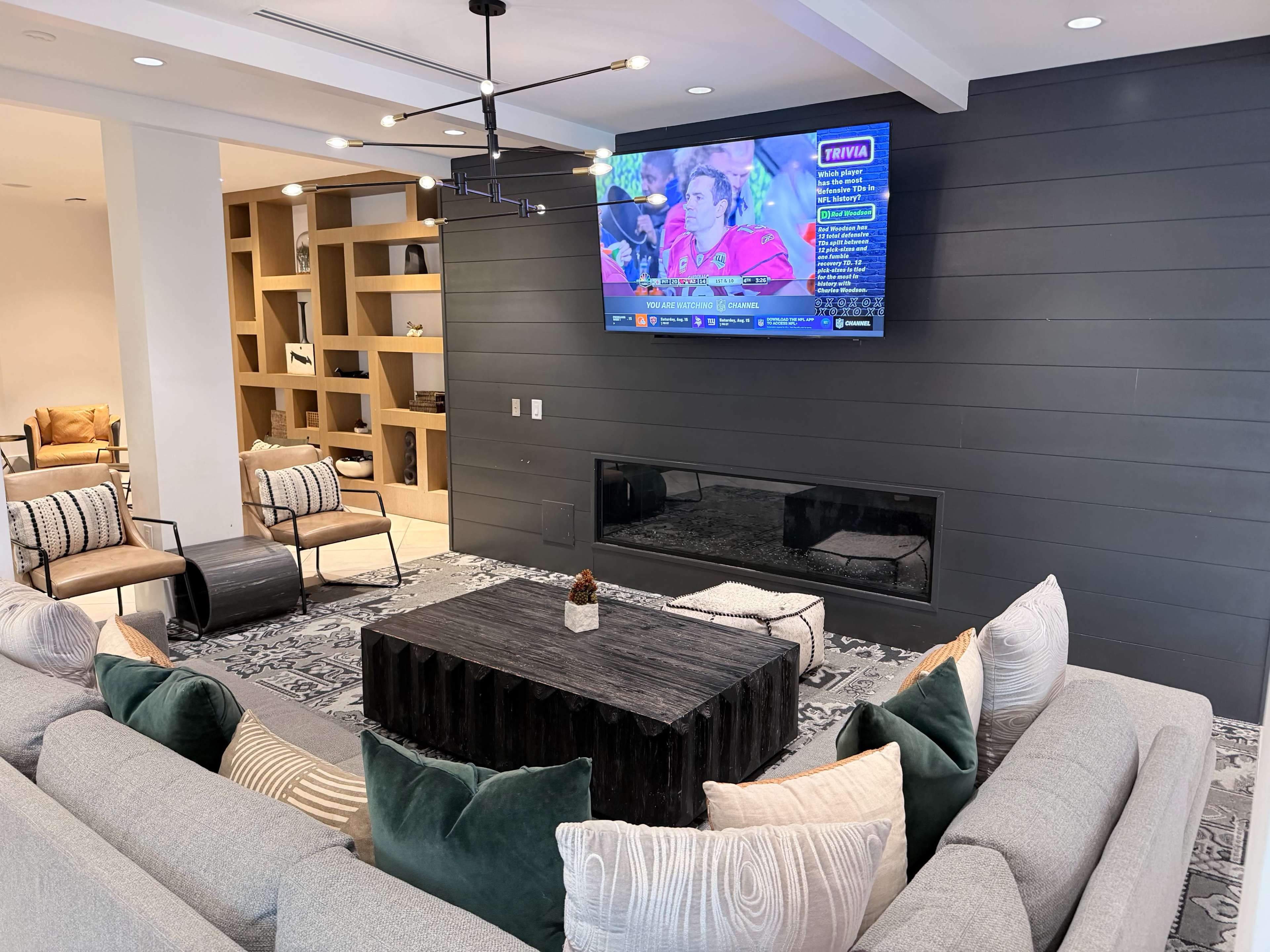 A modern living room features a sectional sofa, a coffee table, and a television mounted on the wall, with a fireplace below and decorative shelves in the background.