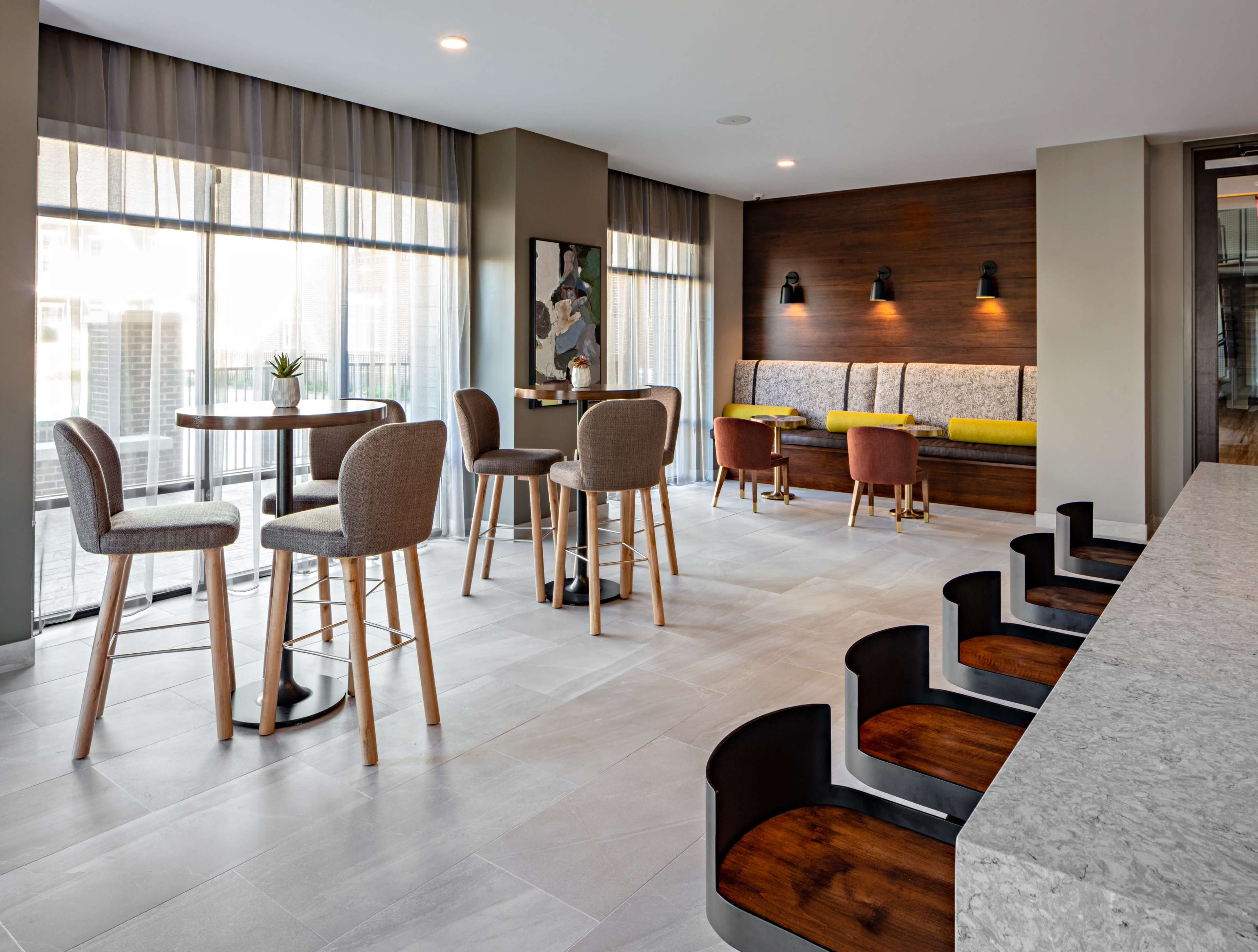A modern interior space featuring round tables and high chairs, a cozy seating area with wooden accents, and large windows allowing natural light to enter.
