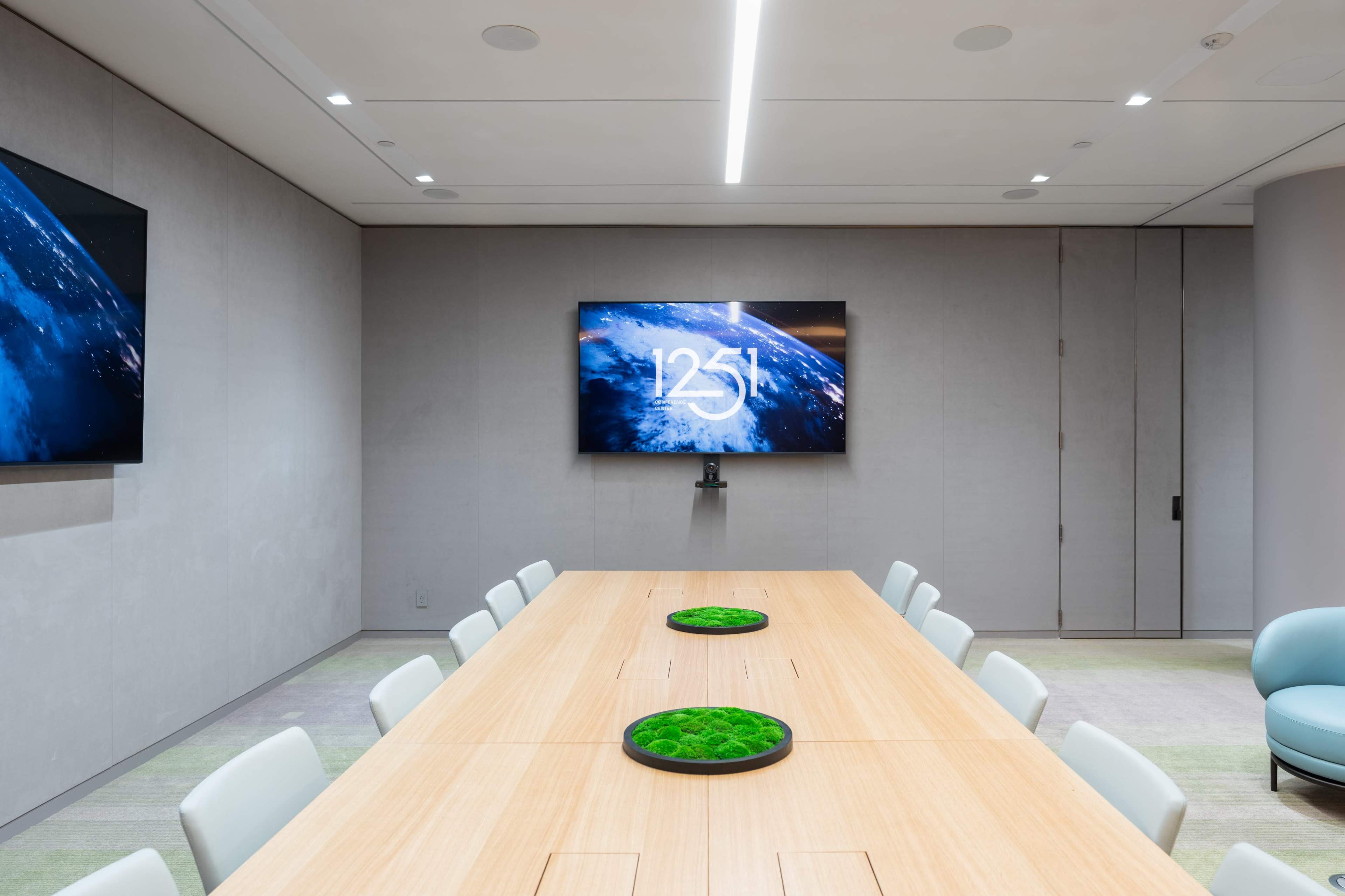 A long wooden conference table is set in a modern meeting room, flanked by green moss displays and large screens displaying a space-related graphic and the number "1251."