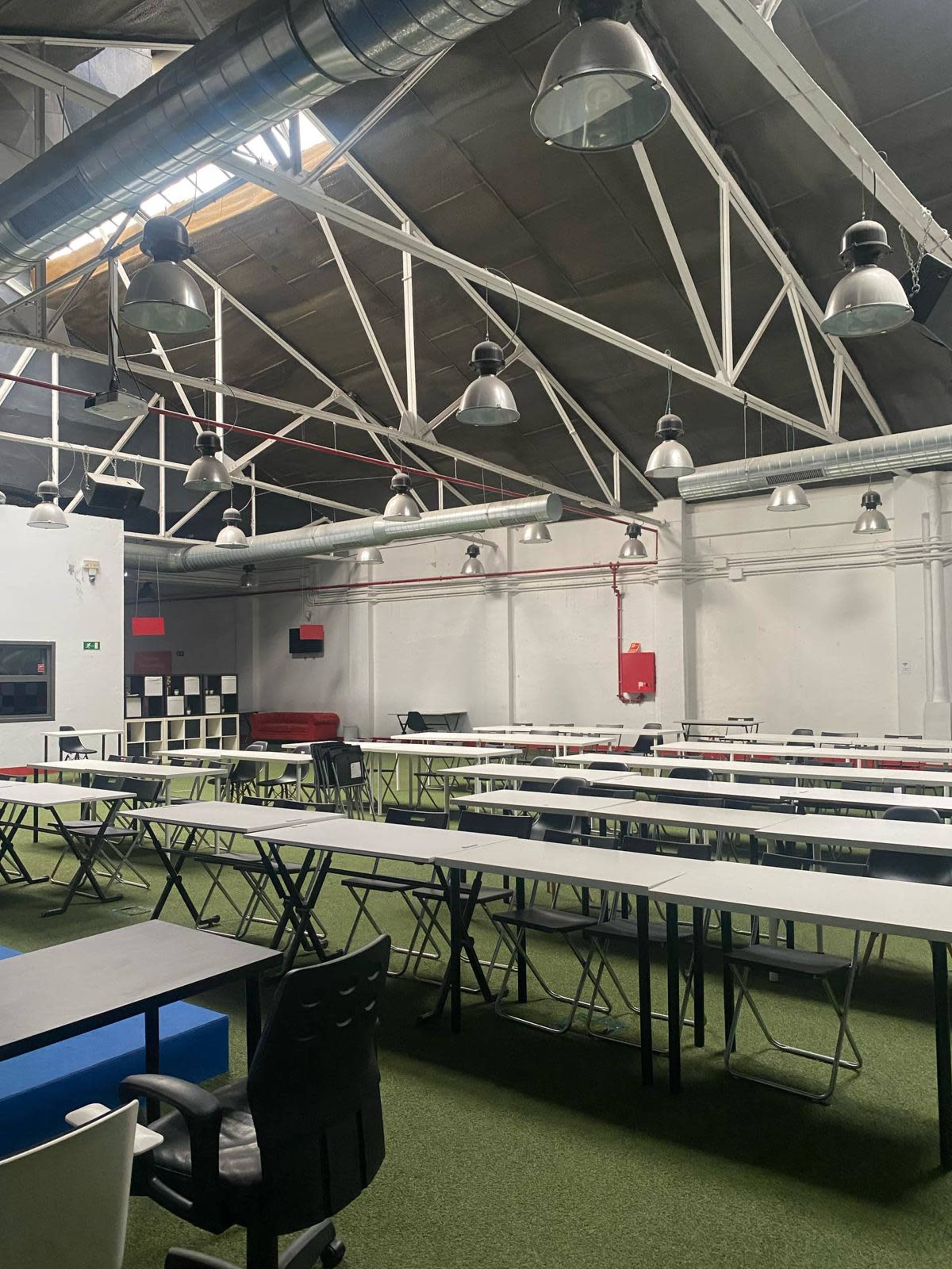 The image shows a spacious, industrial-style room filled with rows of folding tables and chairs set on green artificial grass.