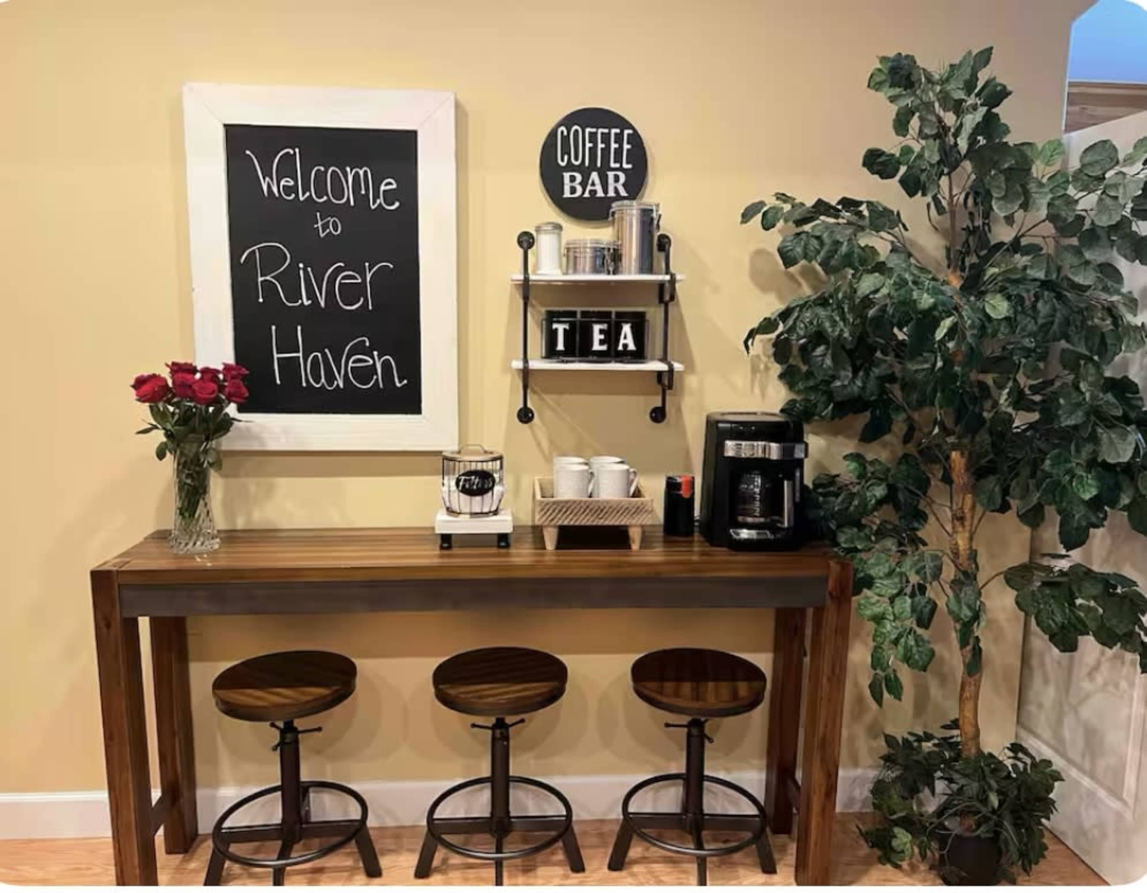 A cozy coffee bar area features a chalkboard welcoming guests to "River Haven," with a small table, coffee maker, and three bar stools beneath a decorative plant.