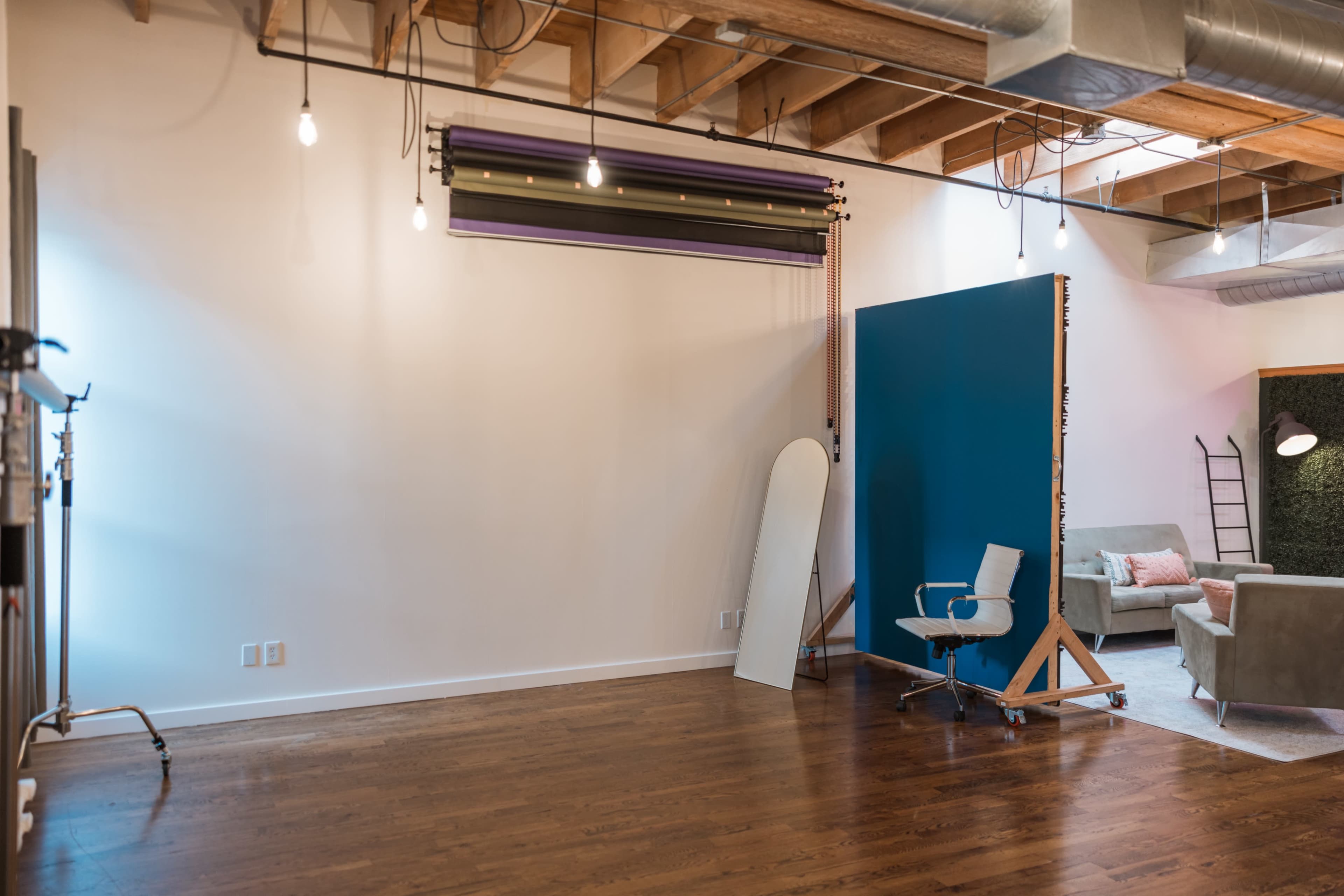 The image shows a spacious, well-lit interior with wooden flooring, featuring a blue backdrop, a chair on wheels, a mirror, and soft seating areas.