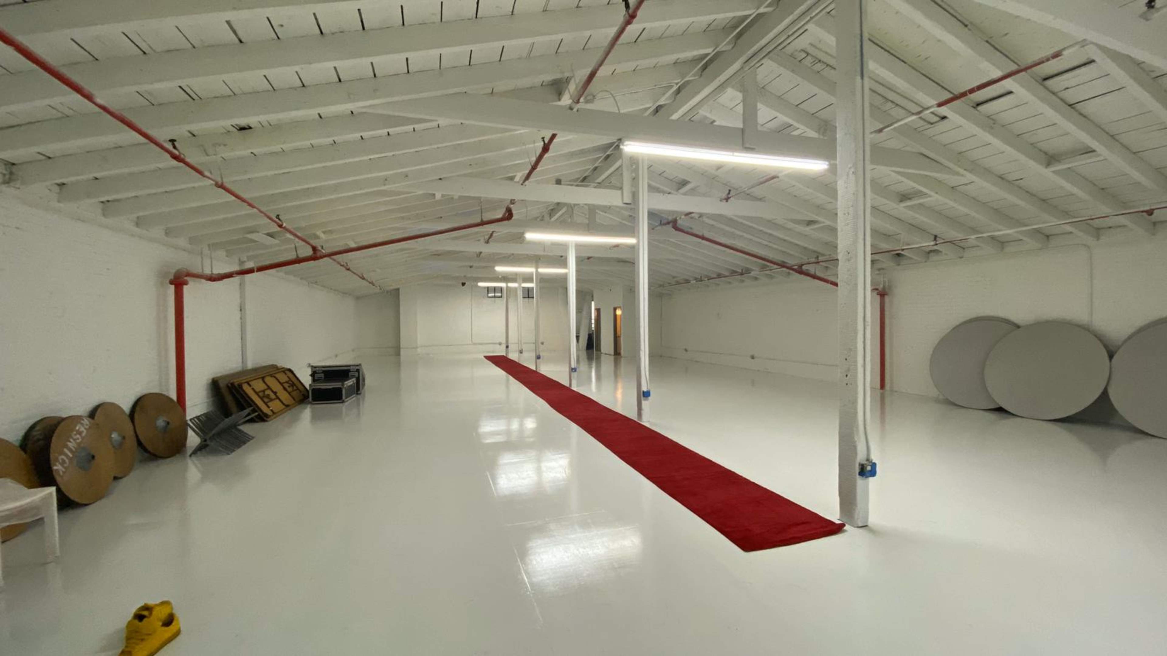 A spacious, white-walled room with high ceilings, a red carpet running down the middle, and various round and rectangular objects leaning against the walls.