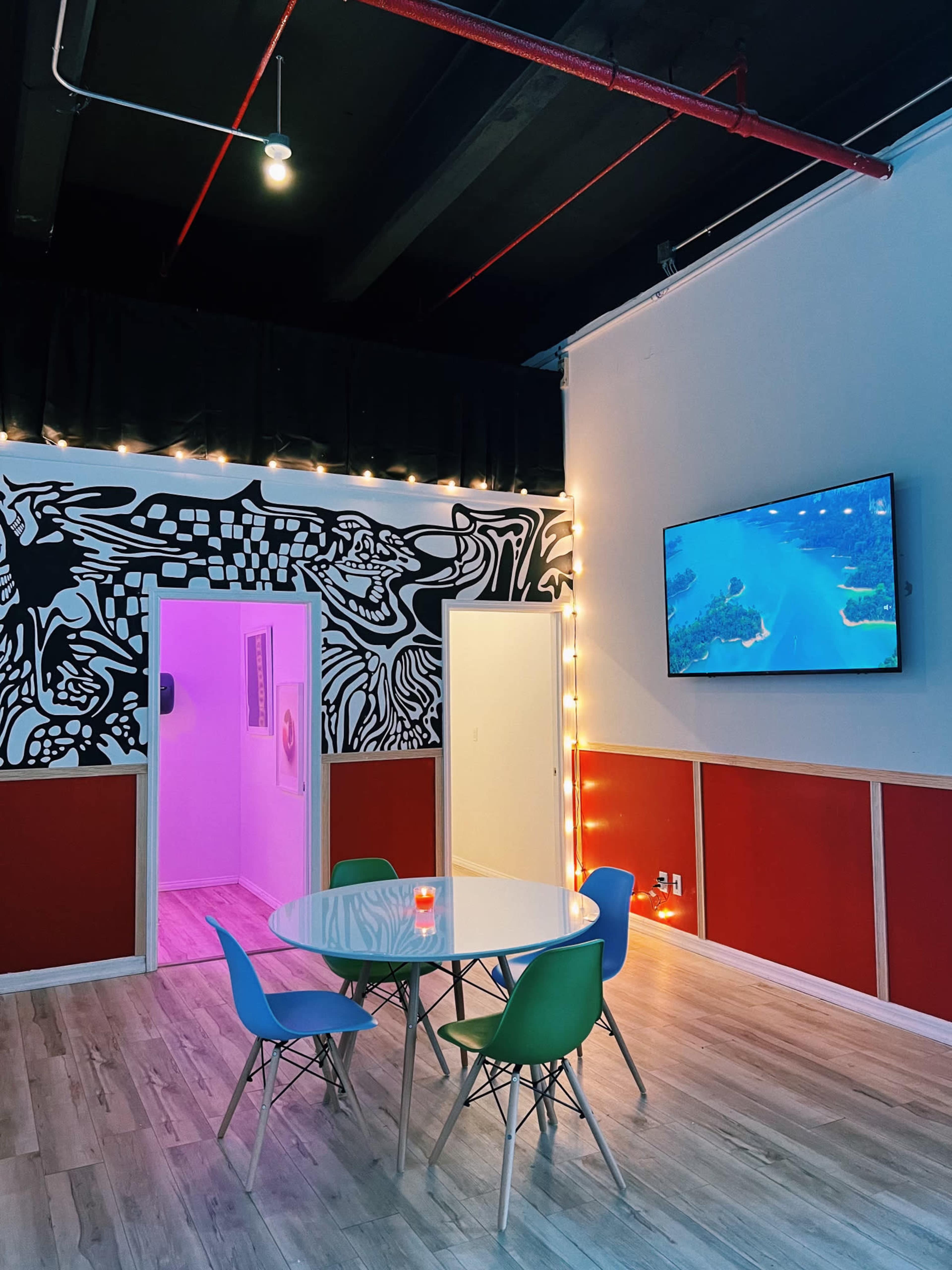 A brightly lit room features a round table surrounded by colorful chairs, with a wall displaying a mural and a television mounted on the opposite wall.