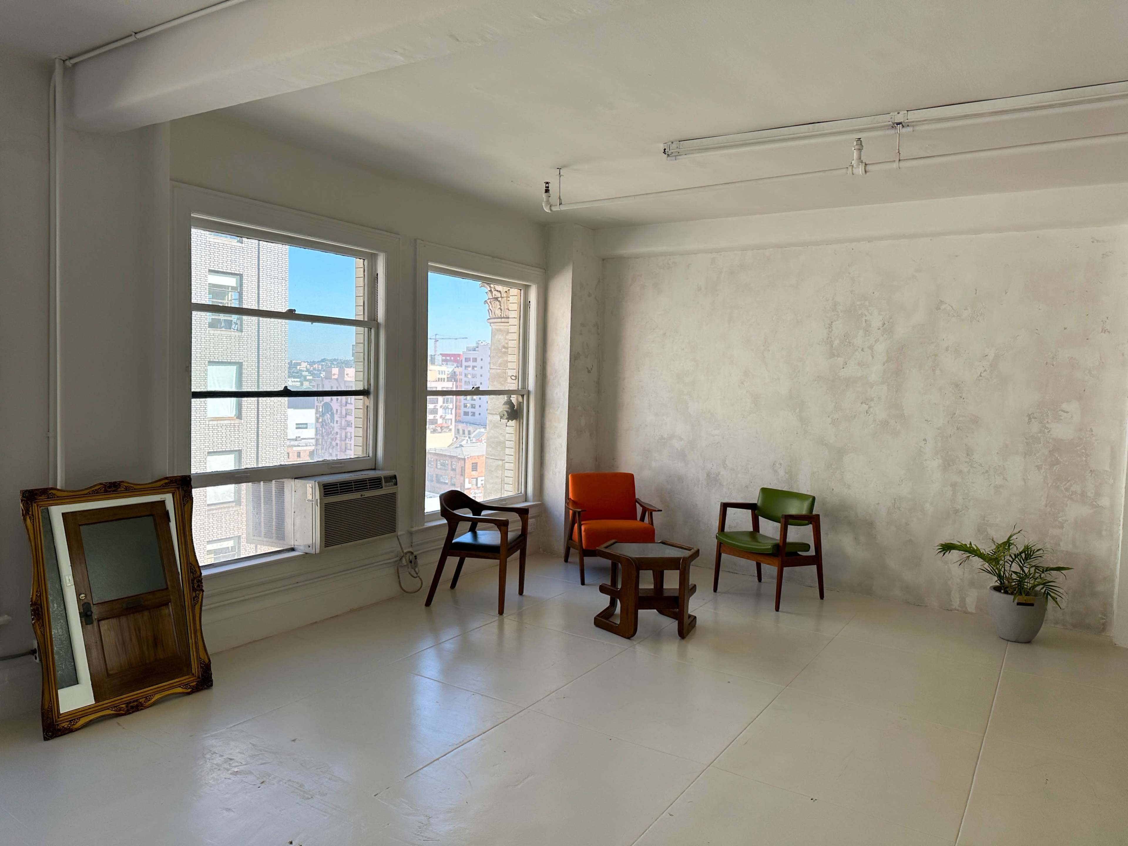 A sparsely furnished room with two colorful chairs, a wooden table, a mirror, and a small plant near a window overlooking a cityscape.