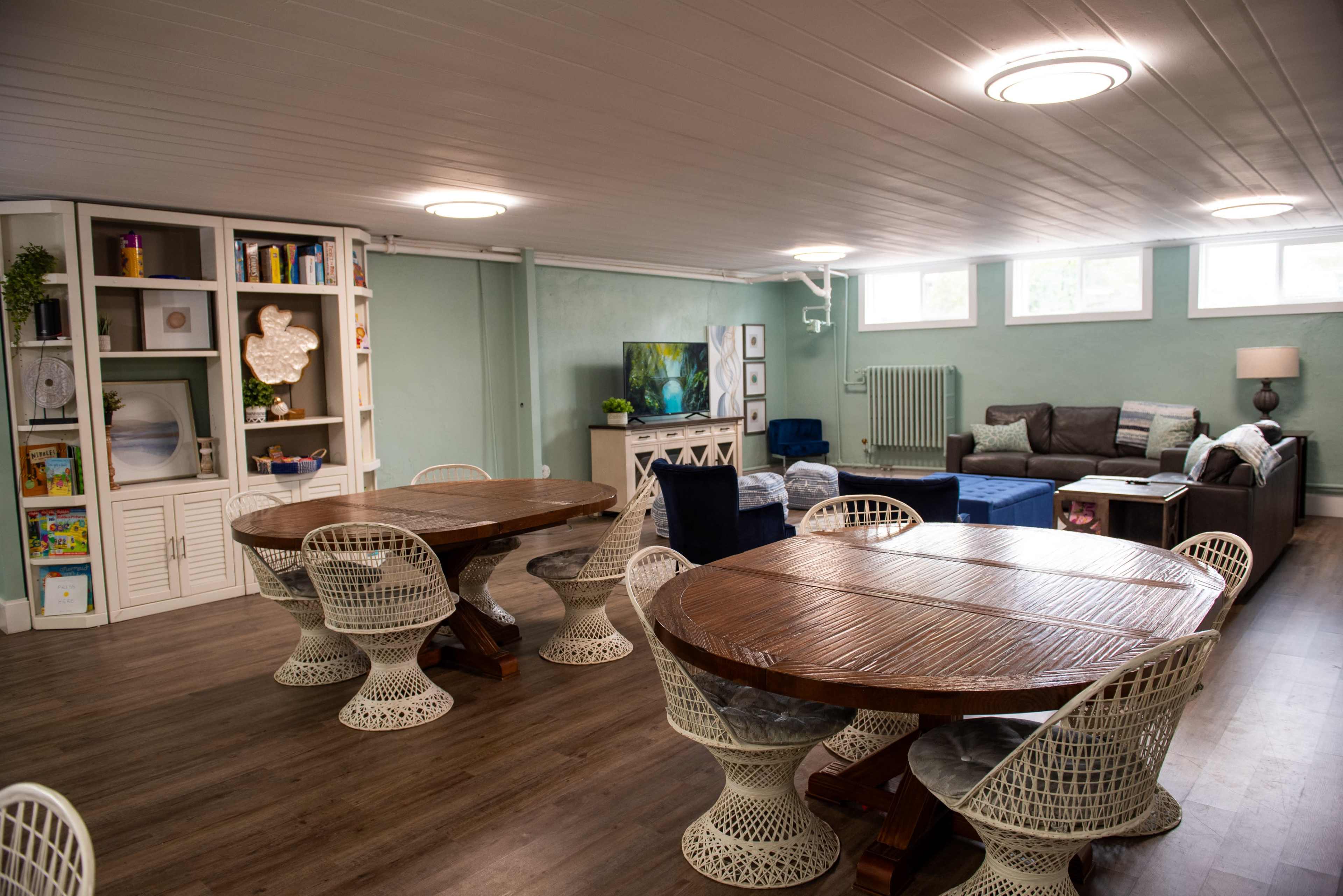 The image shows a spacious basement room with two round dining tables surrounded by woven chairs, a cozy seating area with a couch and a television, and shelves filled with books and decorative items.