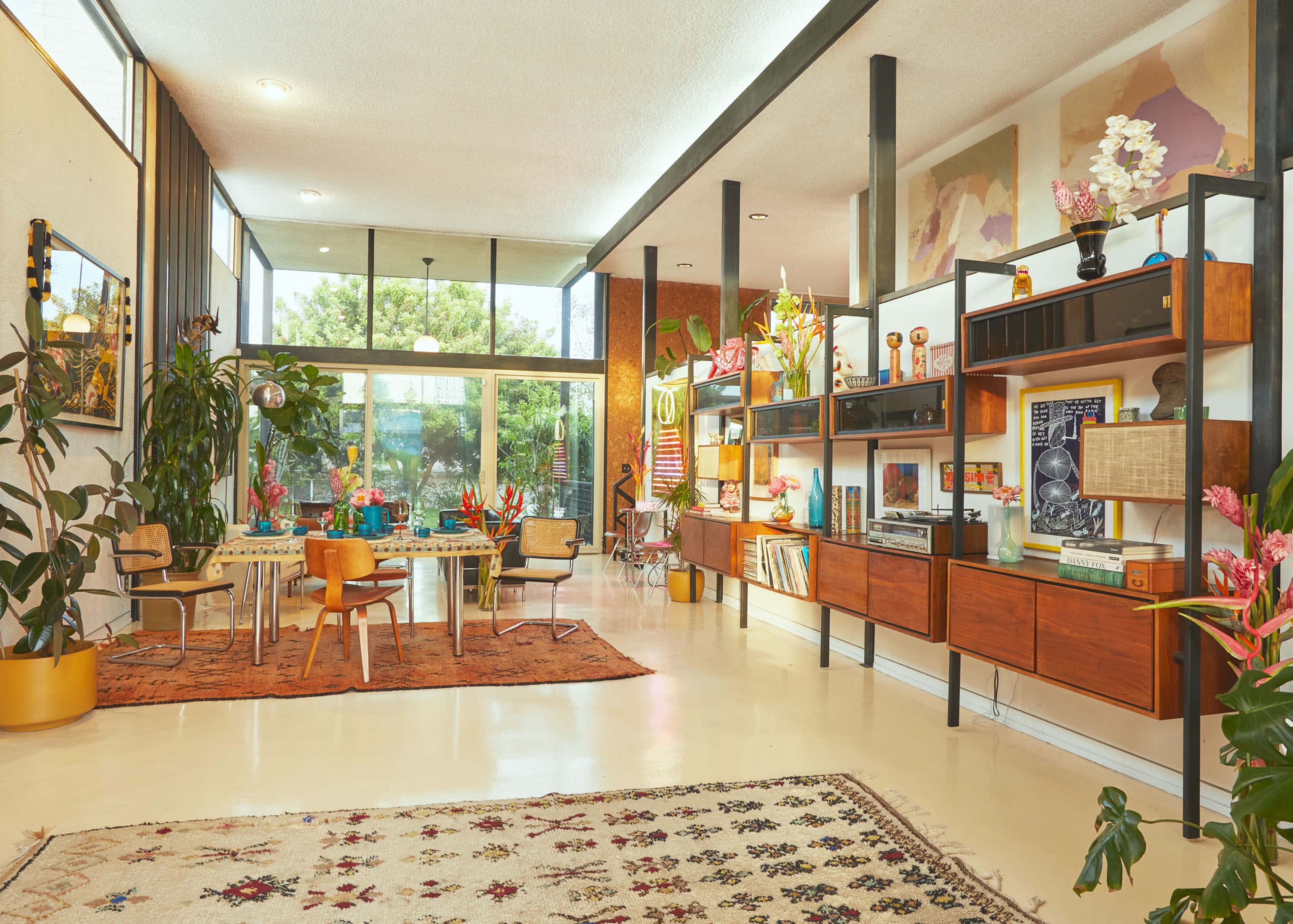 A modern living room features large windows, stylish furniture, and colorful decor, with plants and a dining area complementing the space.
