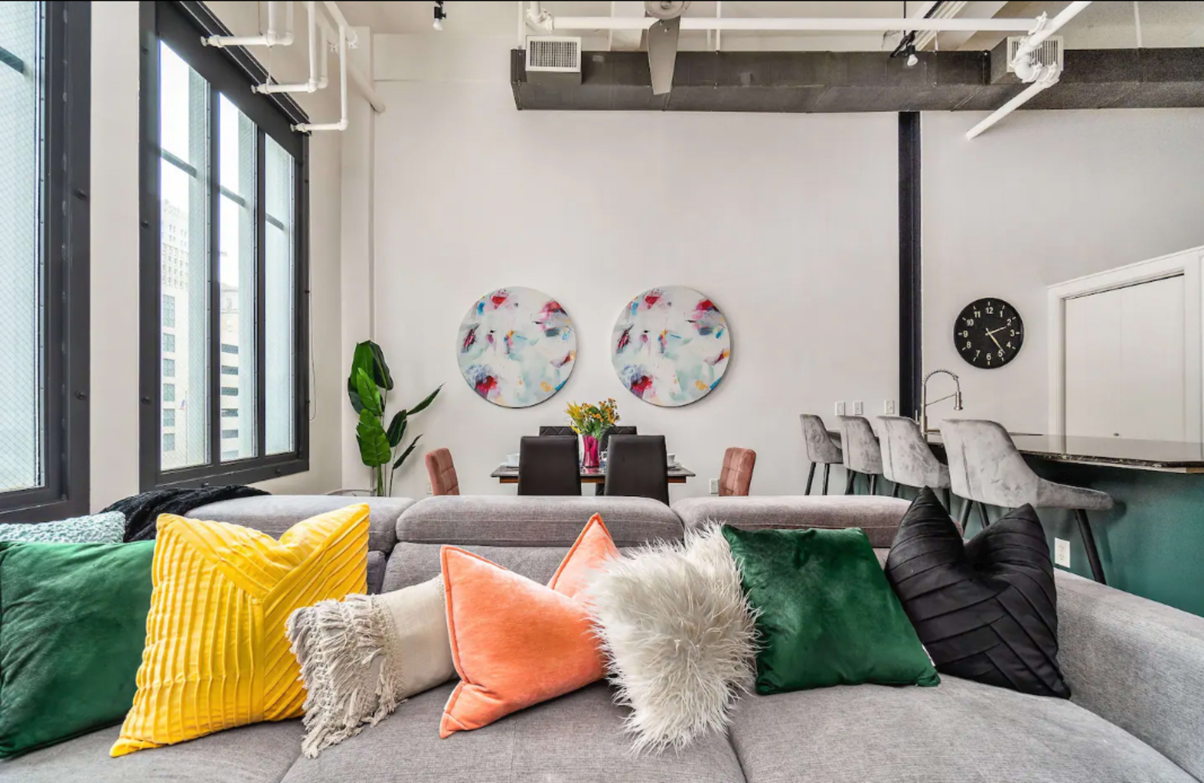 A modern living space featuring a gray sofa adorned with colorful decorative pillows, a dining area in the background, and large windows letting in natural light.