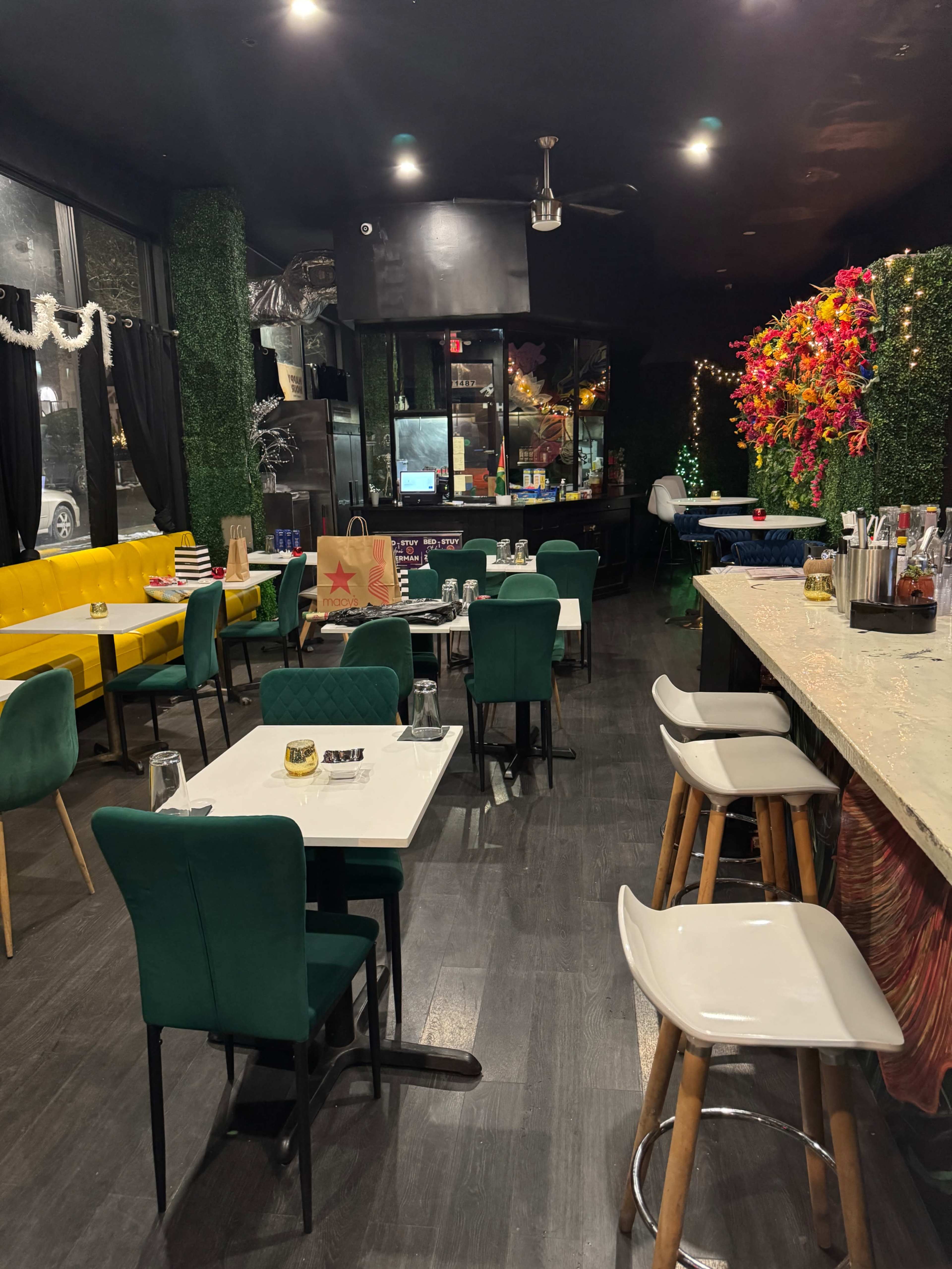 The image shows a restaurant interior featuring green and yellow seating, a bar area, and decorative plants with vibrant floral arrangements.