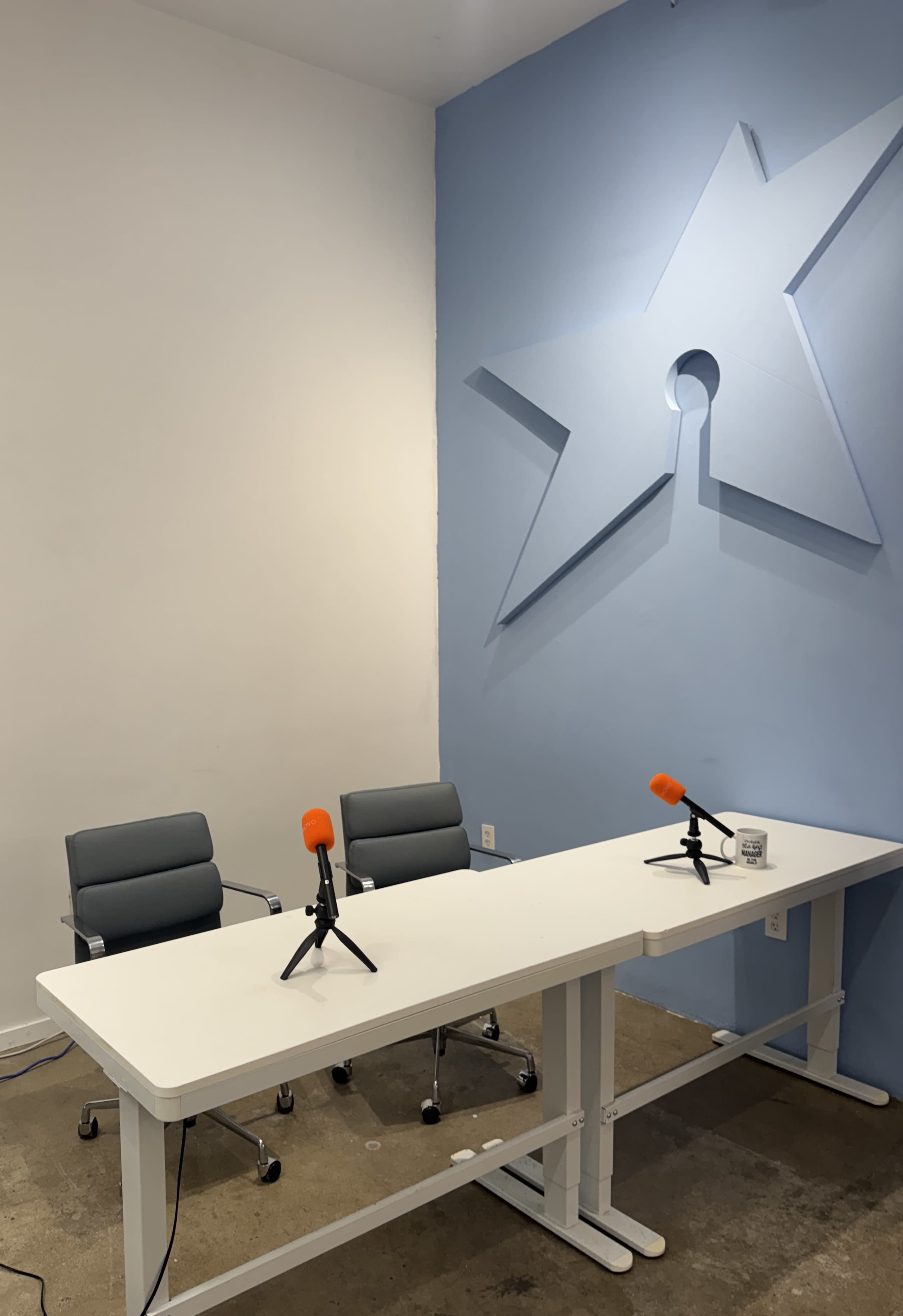 The image shows a minimalist recording or interview setup featuring two gray chairs and a white table with two orange microphones placed on it, against a blue wall with a star design.
