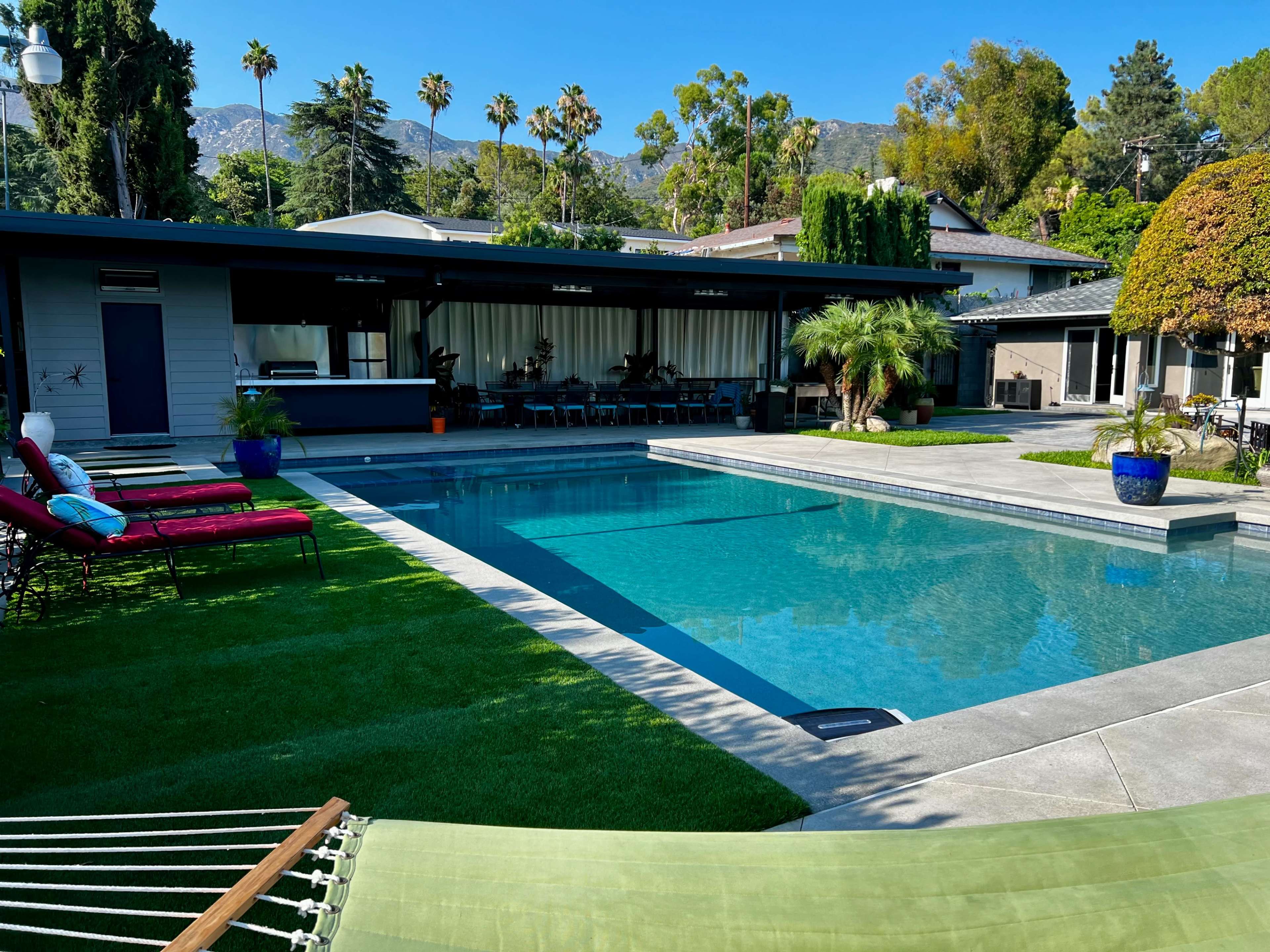 The image features a modern backyard with a swimming pool, lounge chairs, and a covered patio area surrounded by greenery and palm trees.