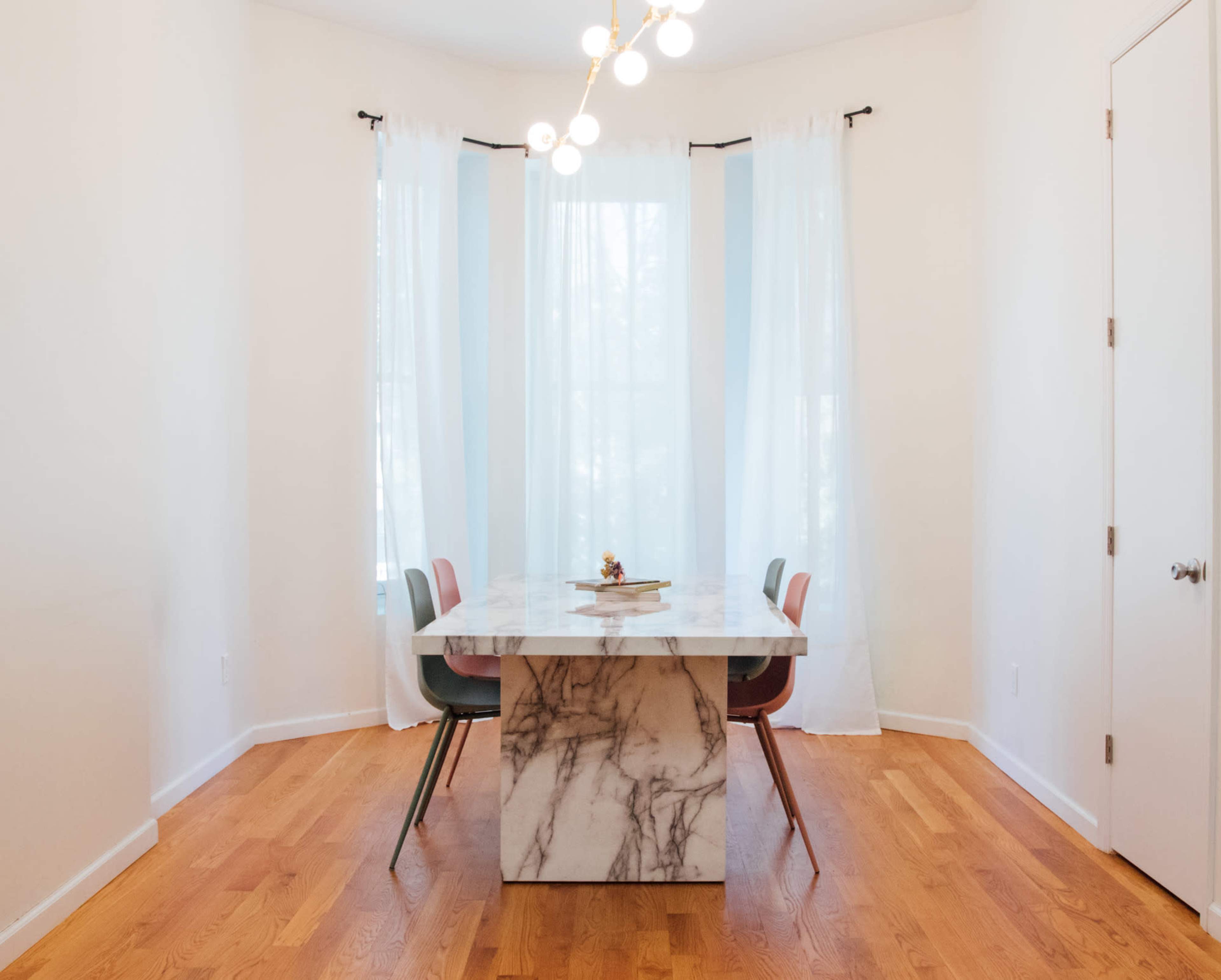 A modern dining area features a marble table surrounded by four colorful chairs, with large windows draped in sheer curtains.