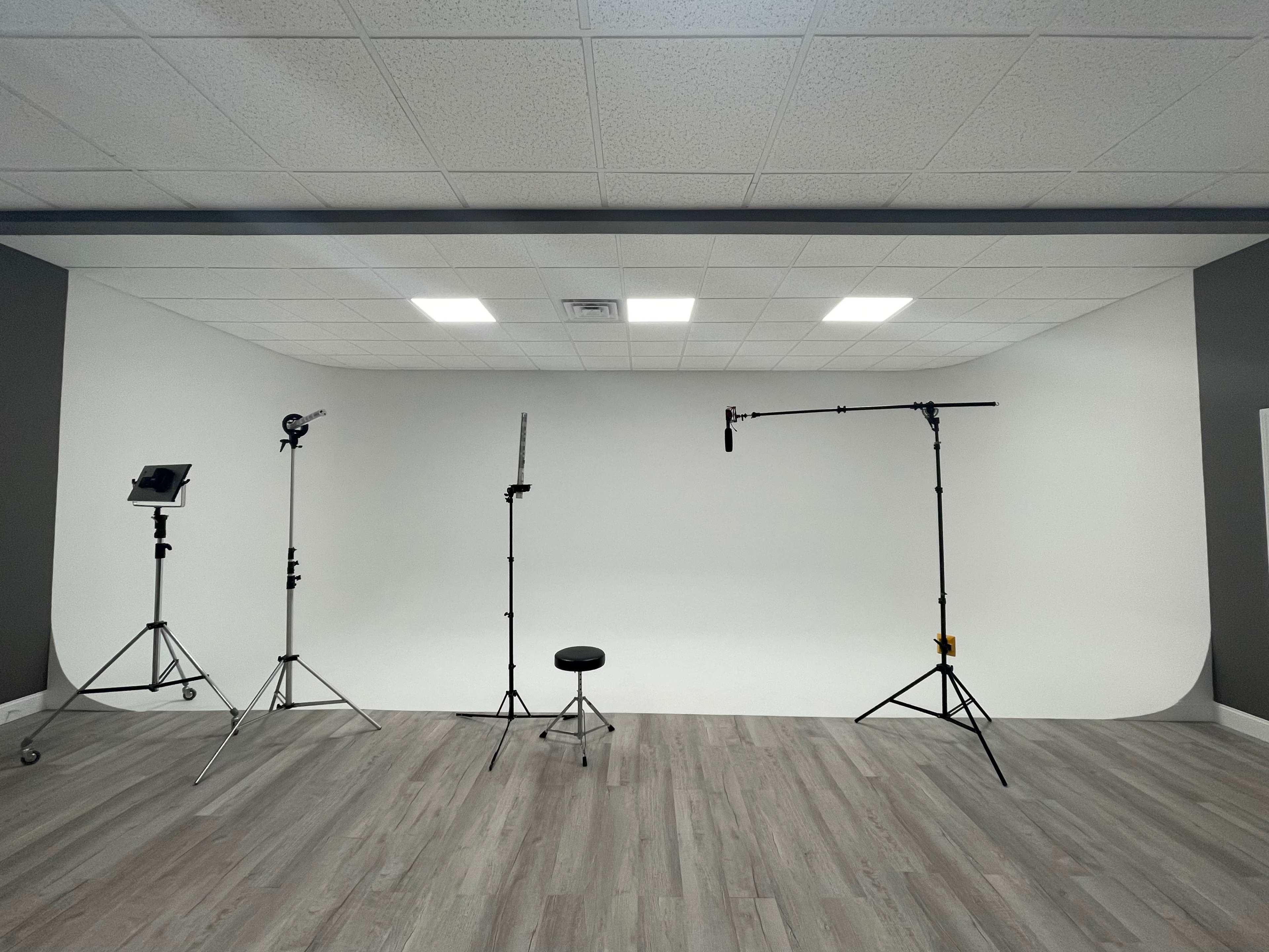 A photography studio with a white backdrop, various lighting equipment, and a single black stool arranged on a wooden floor.