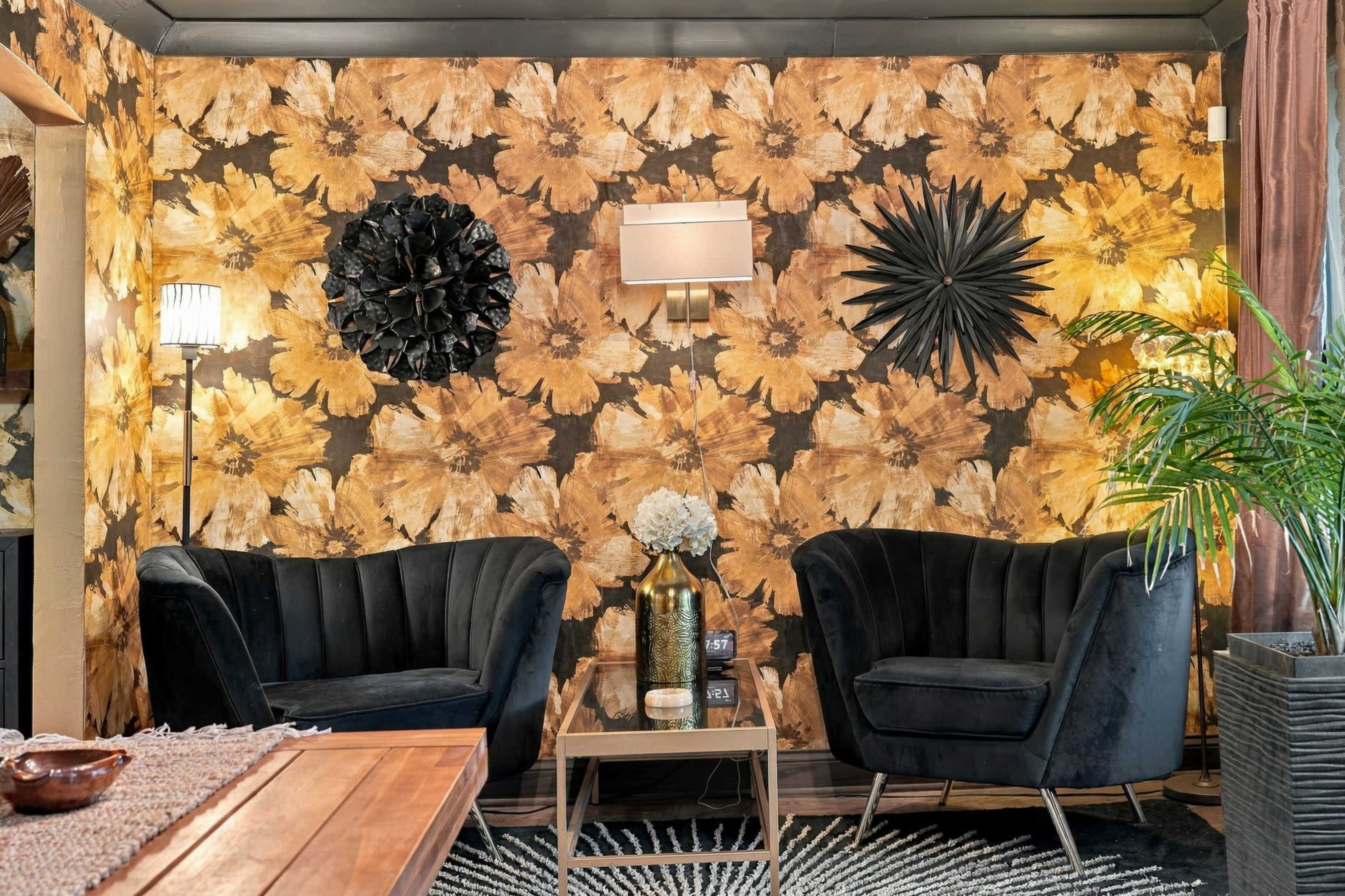 The image features a cozy seating area with two black velvet chairs, a small coffee table, and a decorative wall adorned with large floral-patterned wallpaper.