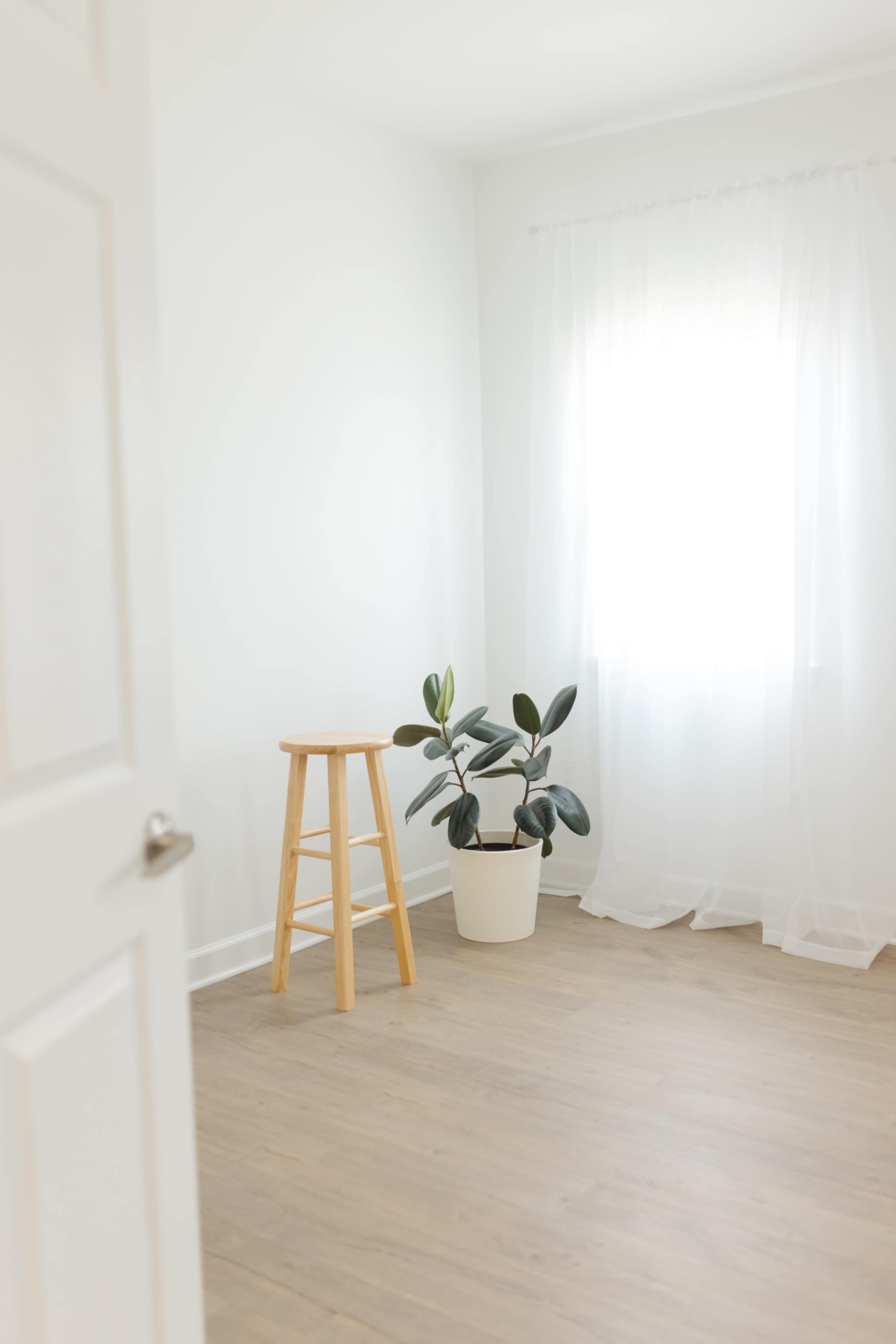 The image shows a bright, minimalistic room with a wooden stool next to a potted plant, illuminated by natural light coming through a sheer curtain.