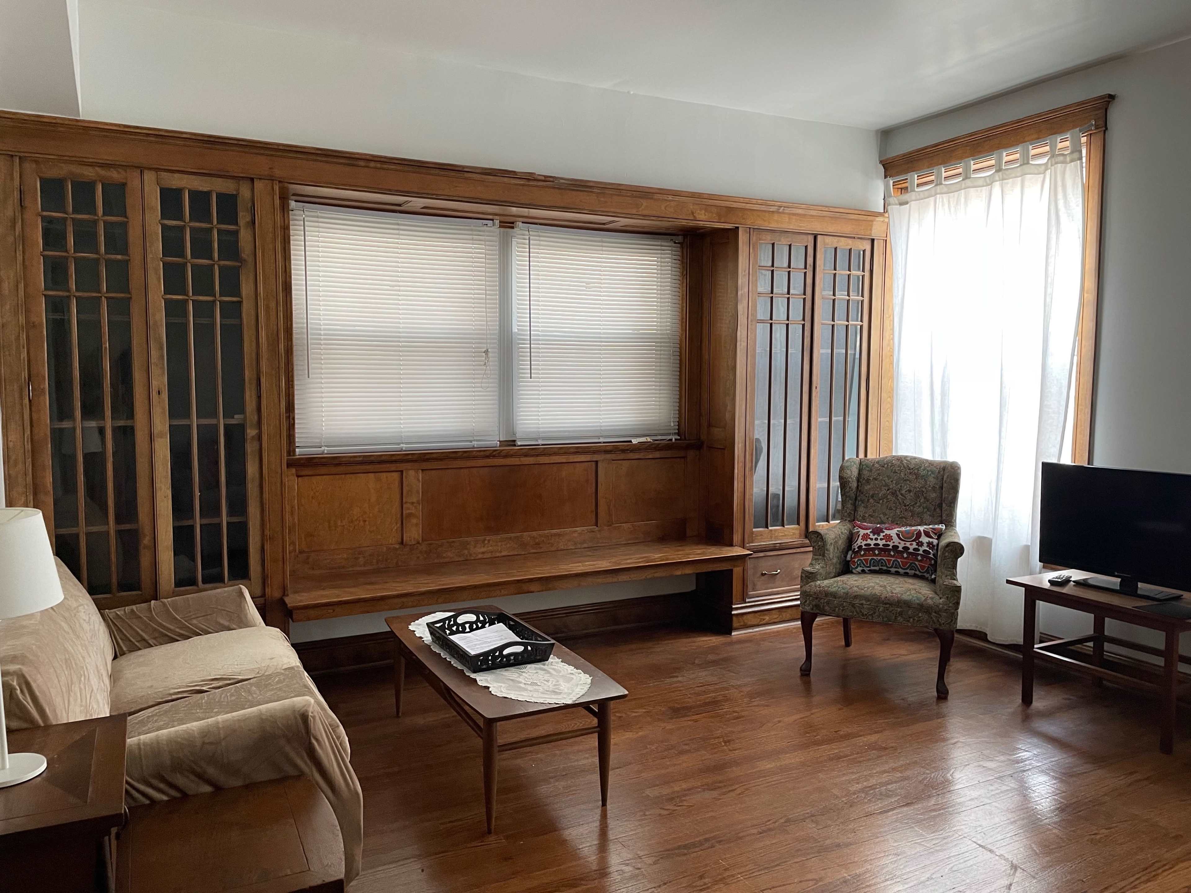 A living room with wooden cabinetry, a light-colored sofa, a patterned chair, a small television on a stand, and a window with sheer curtains.