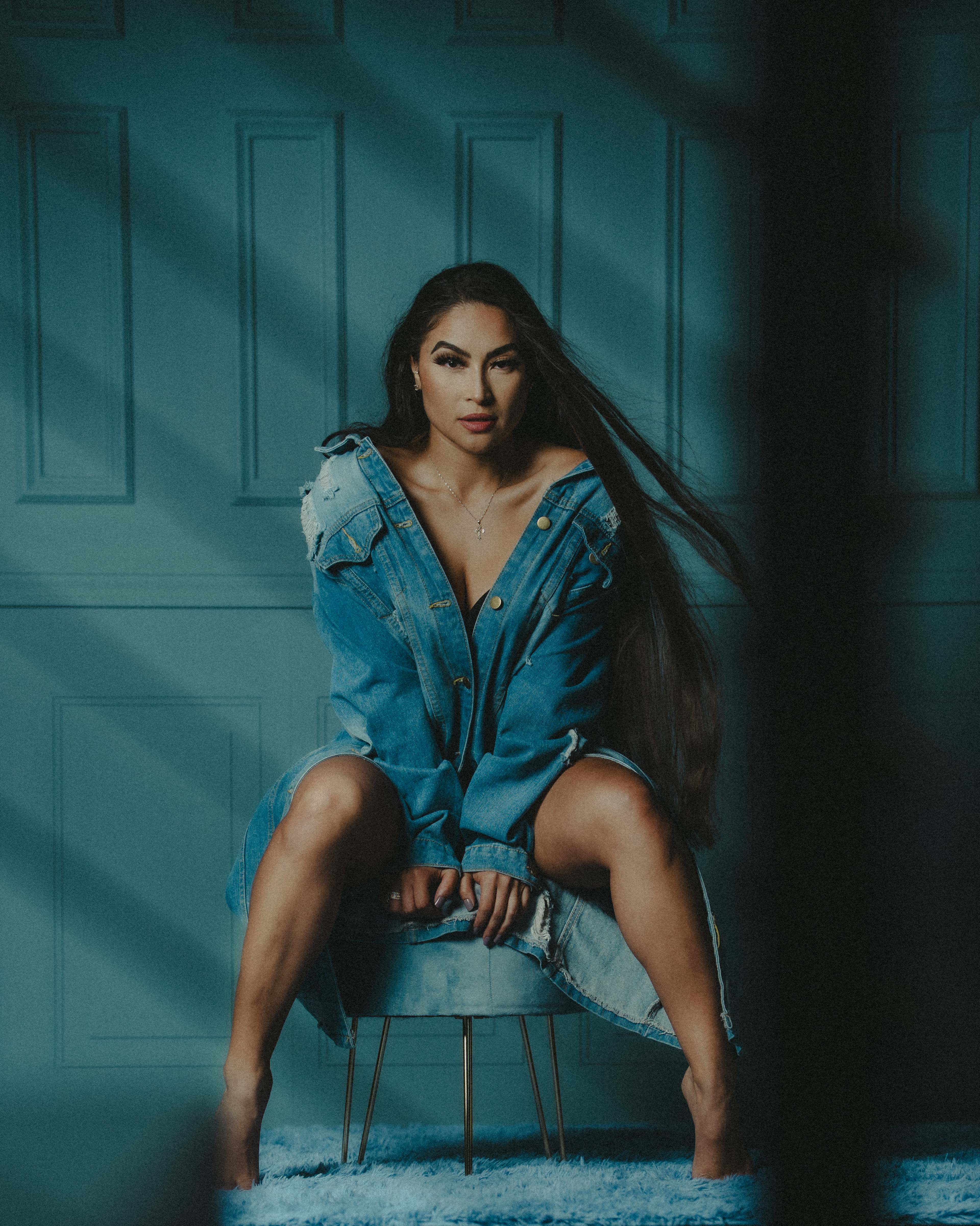 A woman with long hair sits on a small stool, wearing an oversized denim jacket in a room with blue walls.