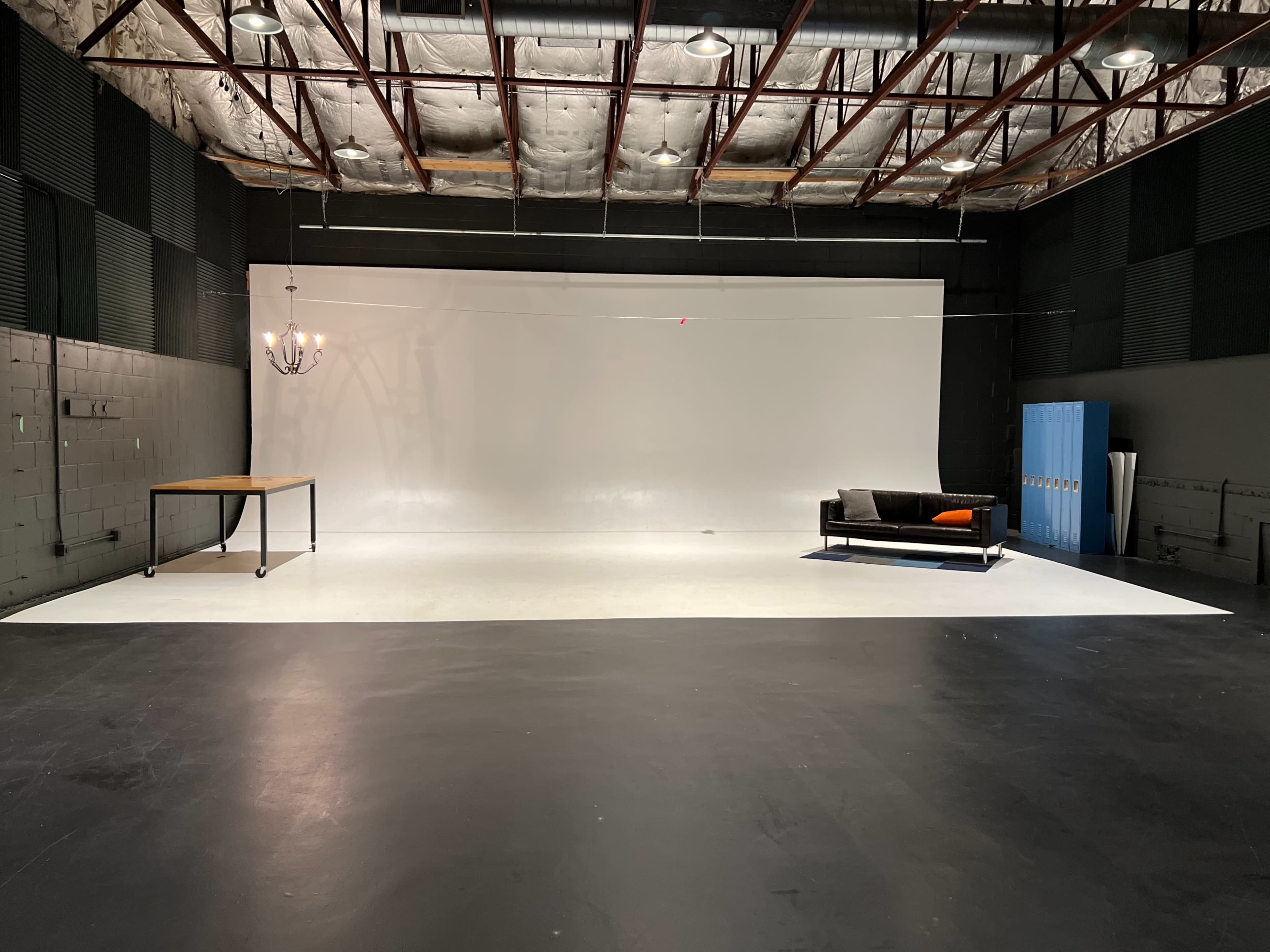 A spacious studio with a white backdrop, a wooden table, a black sofa, and a blue cabinet against the wall.