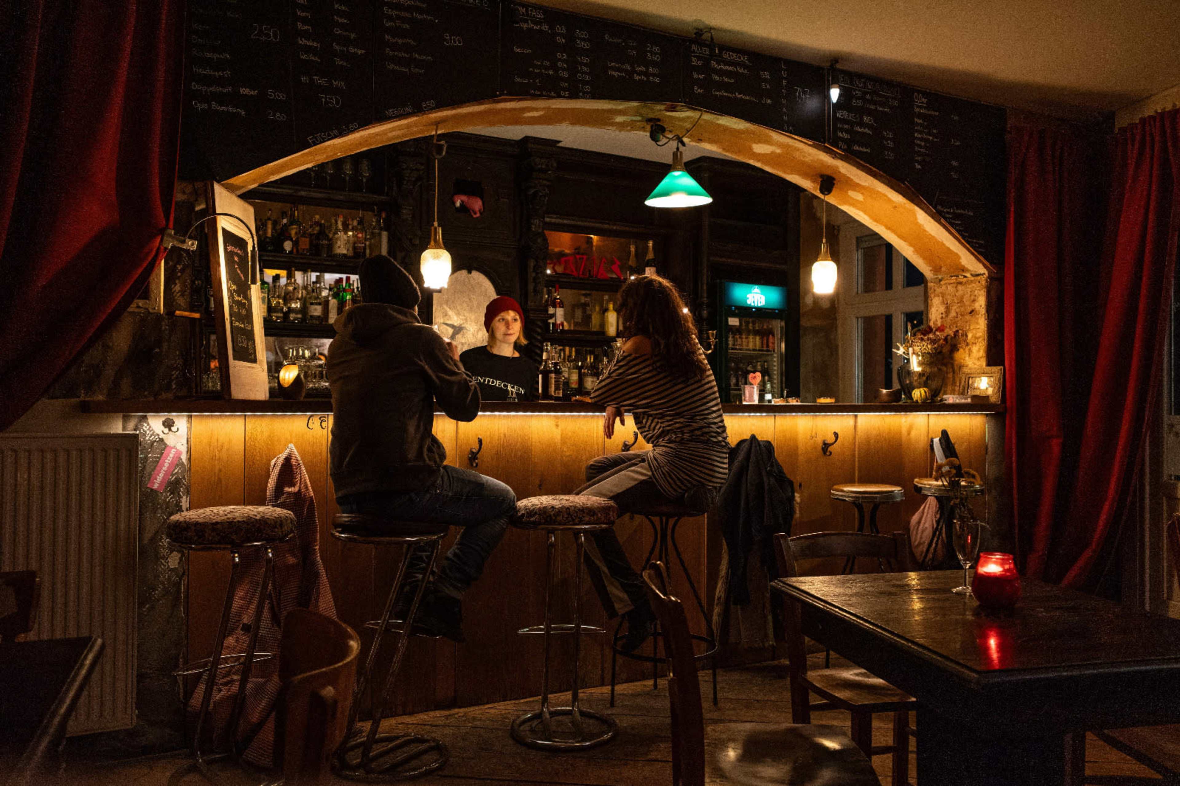 Two patrons sit at a bar counter, conversing with a bartender in a dimly lit establishment adorned with red curtains and a chalkboard menu.