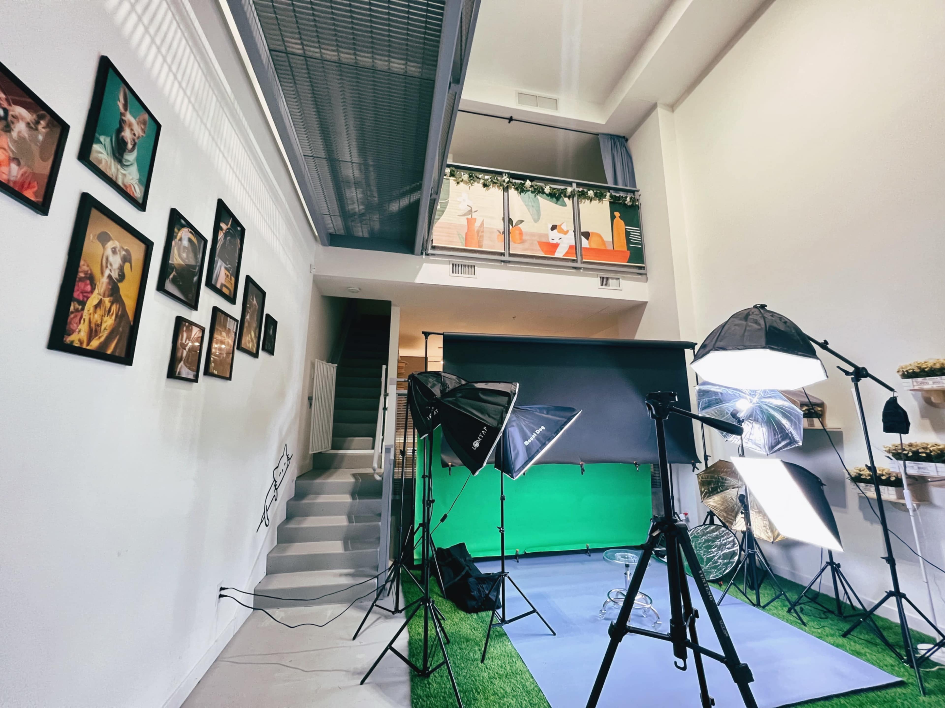 A spacious studio setup with professional lighting and a green screen, surrounded by framed photographs on the walls and a staircase leading to an upper level.