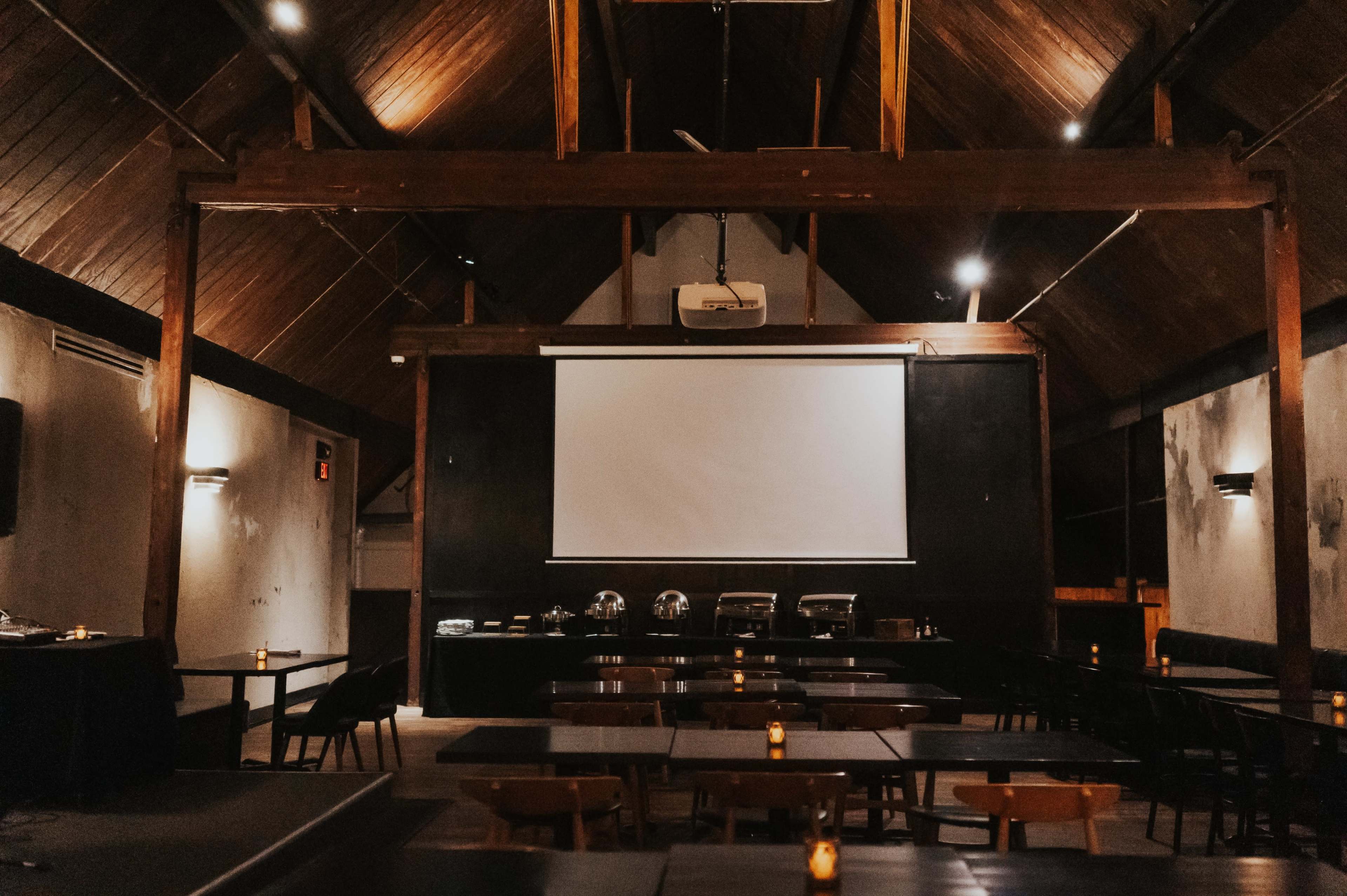 A spacious, dimly lit indoor setting features wooden beams, rows of tables, and a large projection screen at the front.