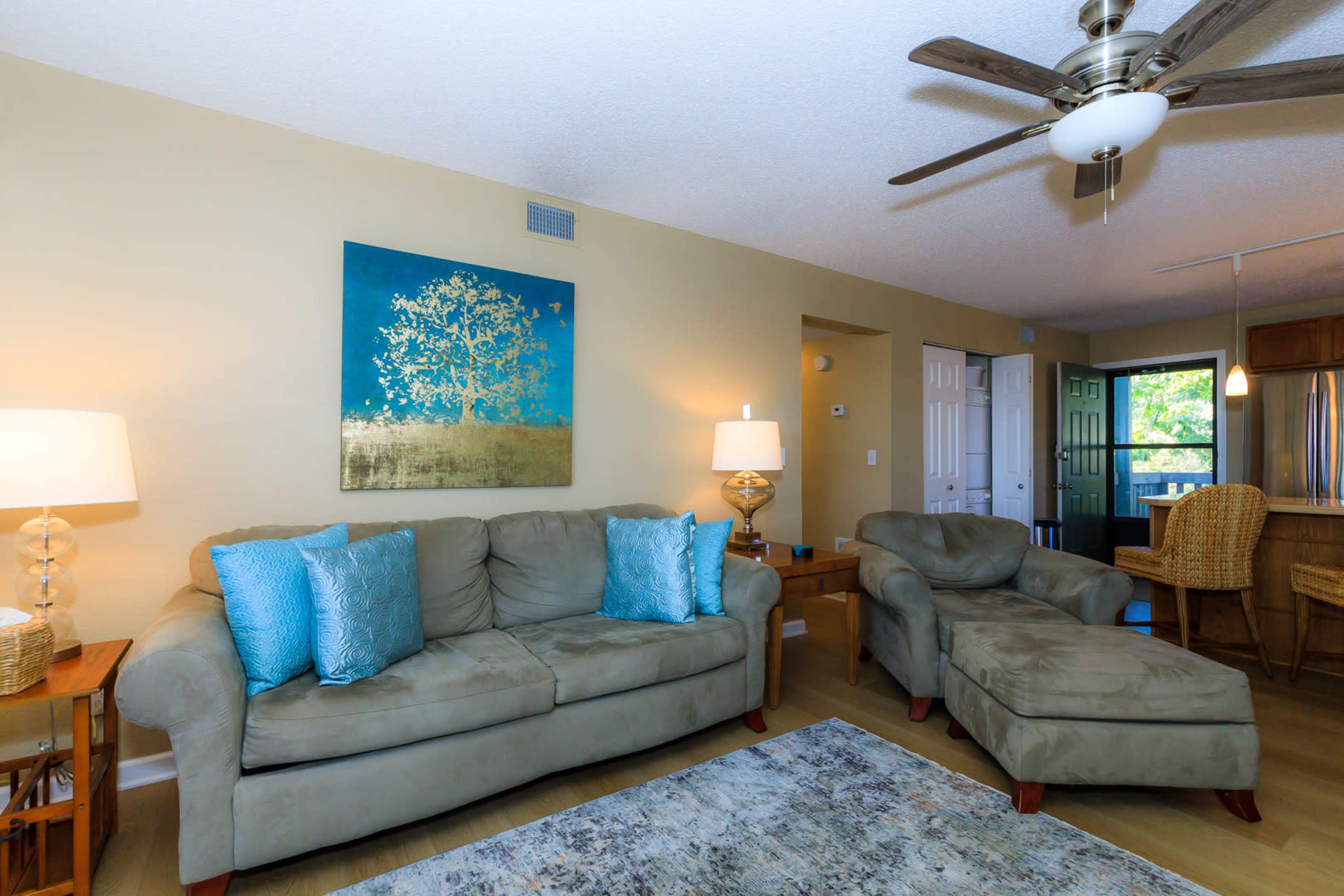 A living room with a light-colored sofa and chaise lounge, a lamp, a decorative wall art featuring a tree, and a view of an adjacent room with a kitchen.