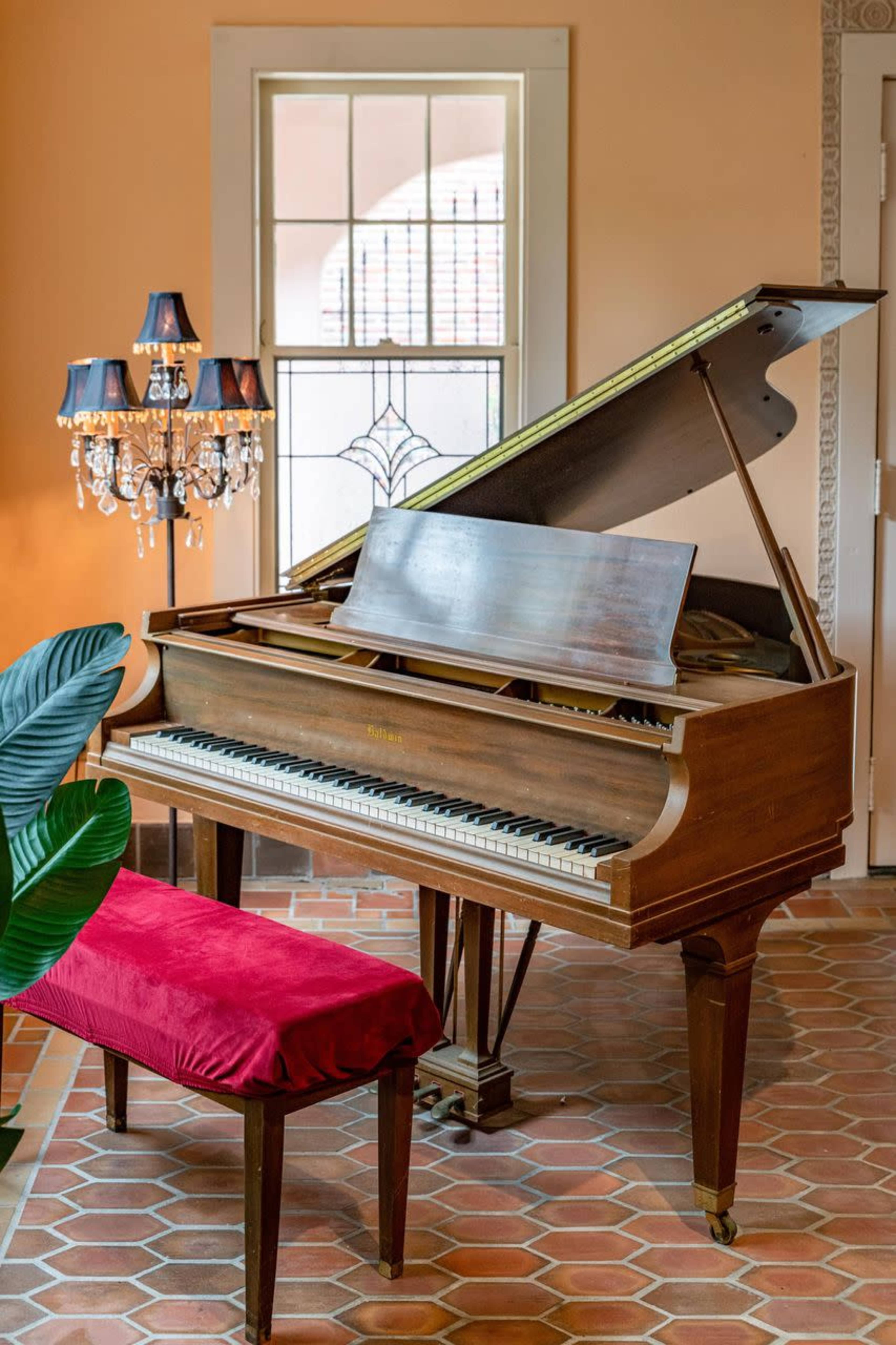 A grand piano with a raised lid sits beside a velvet-covered bench and a decorative lamp in a sunlit room.