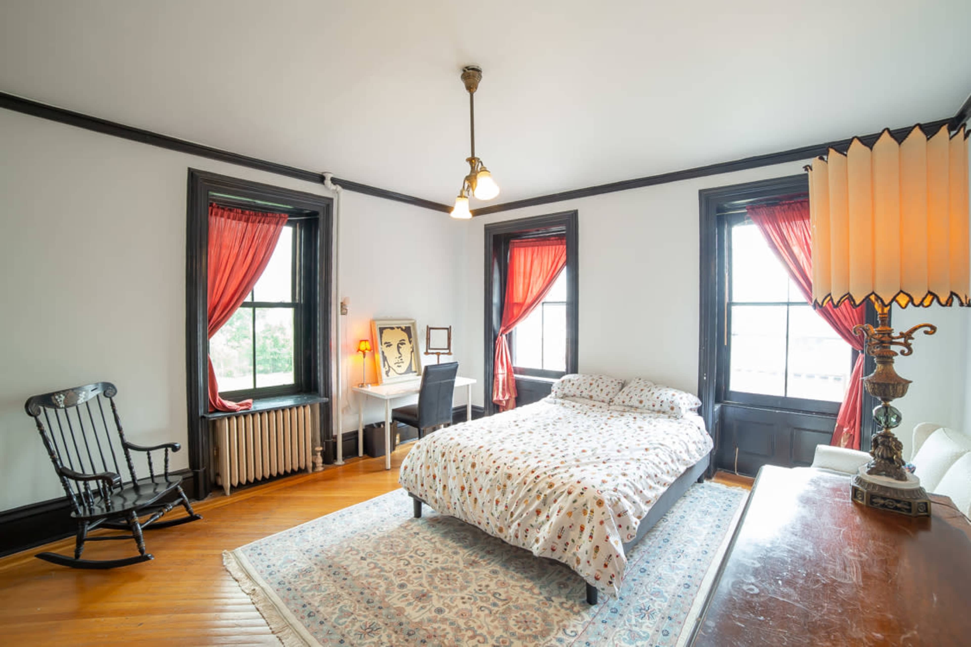 A bedroom with a bed covered in a polka-dotted duvet, a rocking chair, a desk, and large windows with red curtains.
