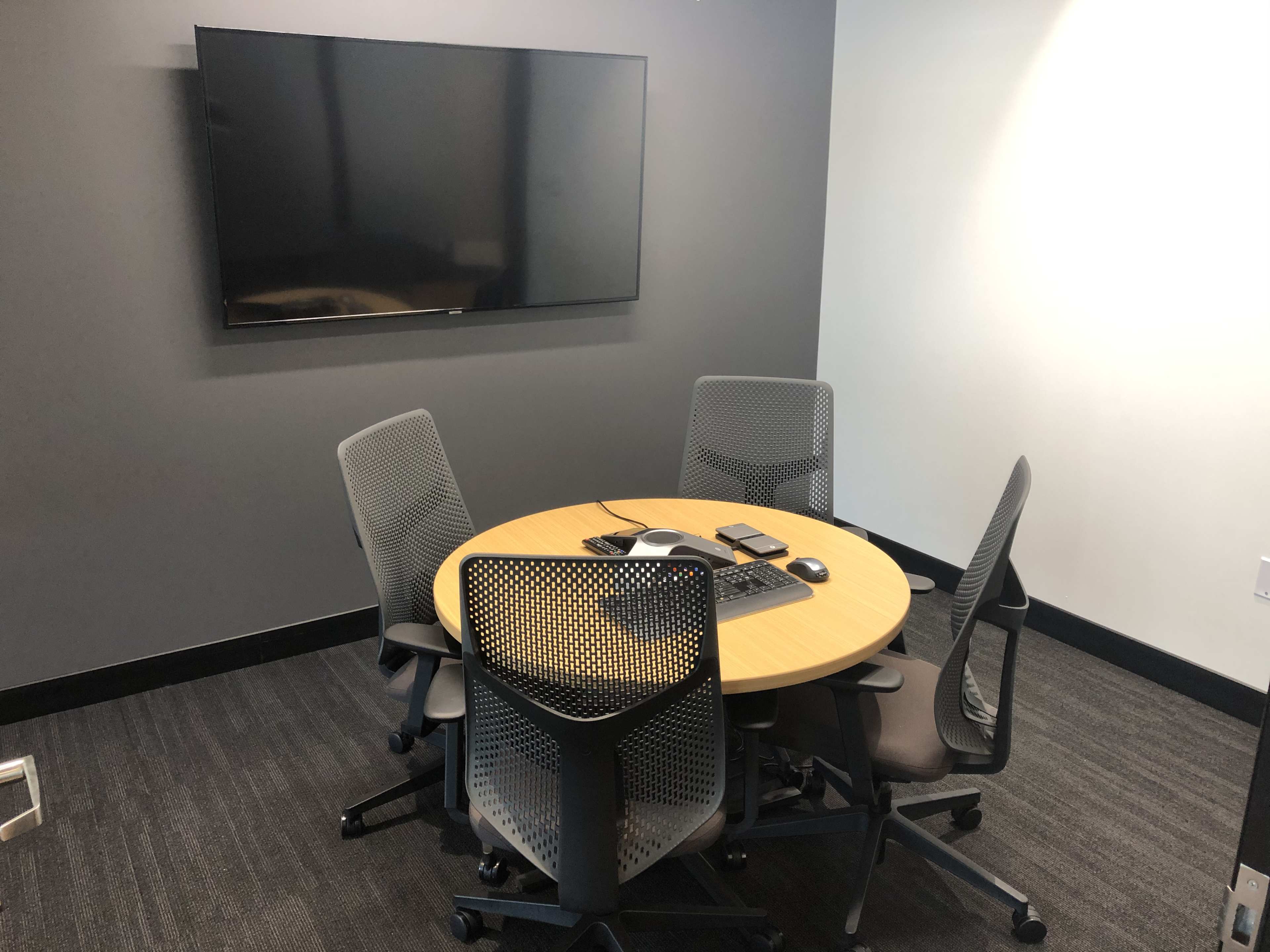 A small conference room features a round table surrounded by four mesh chairs and a wall-mounted television.
