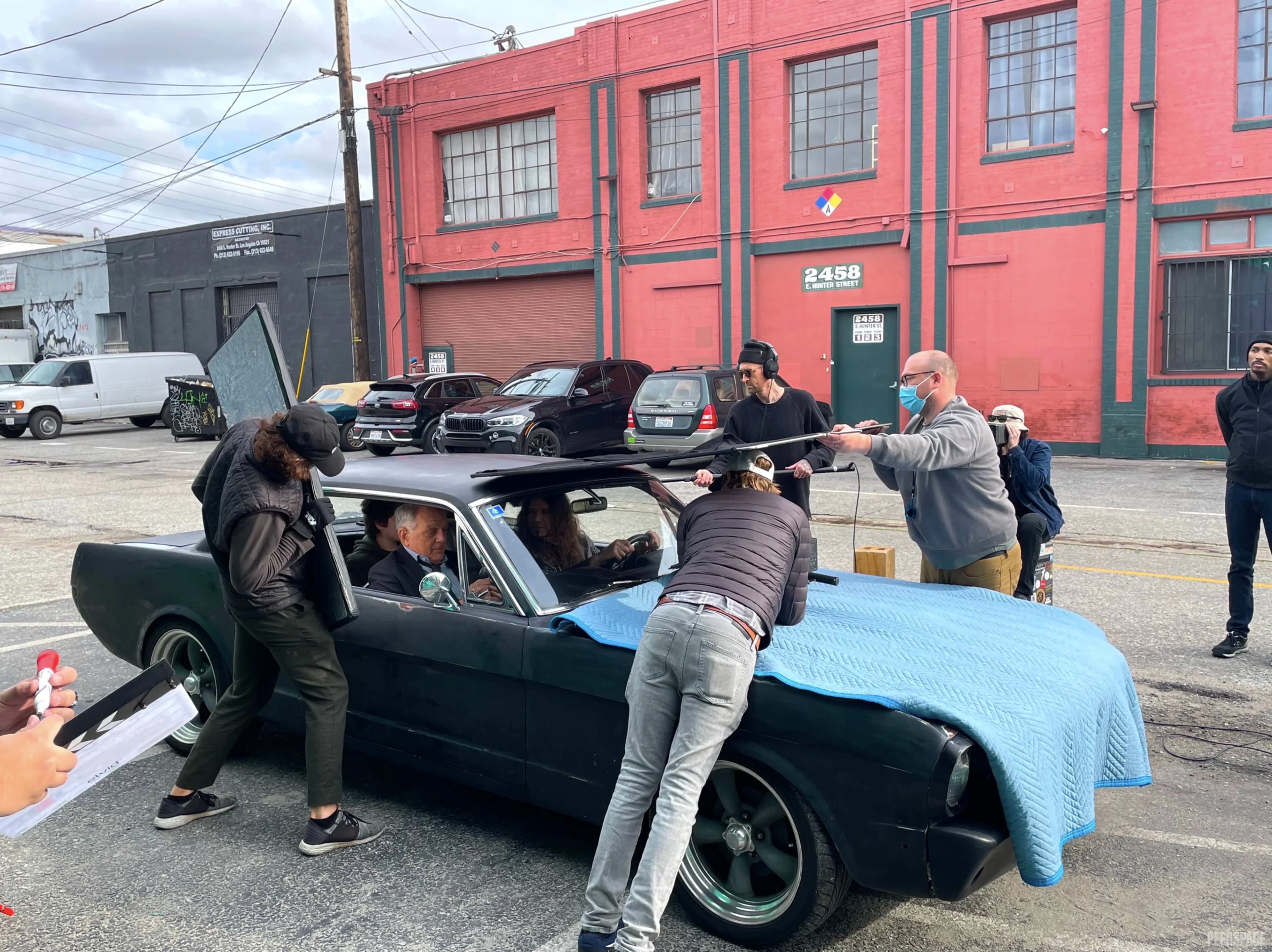 A group of people is filming a scene involving a black car in an urban setting with colorful buildings in the background.