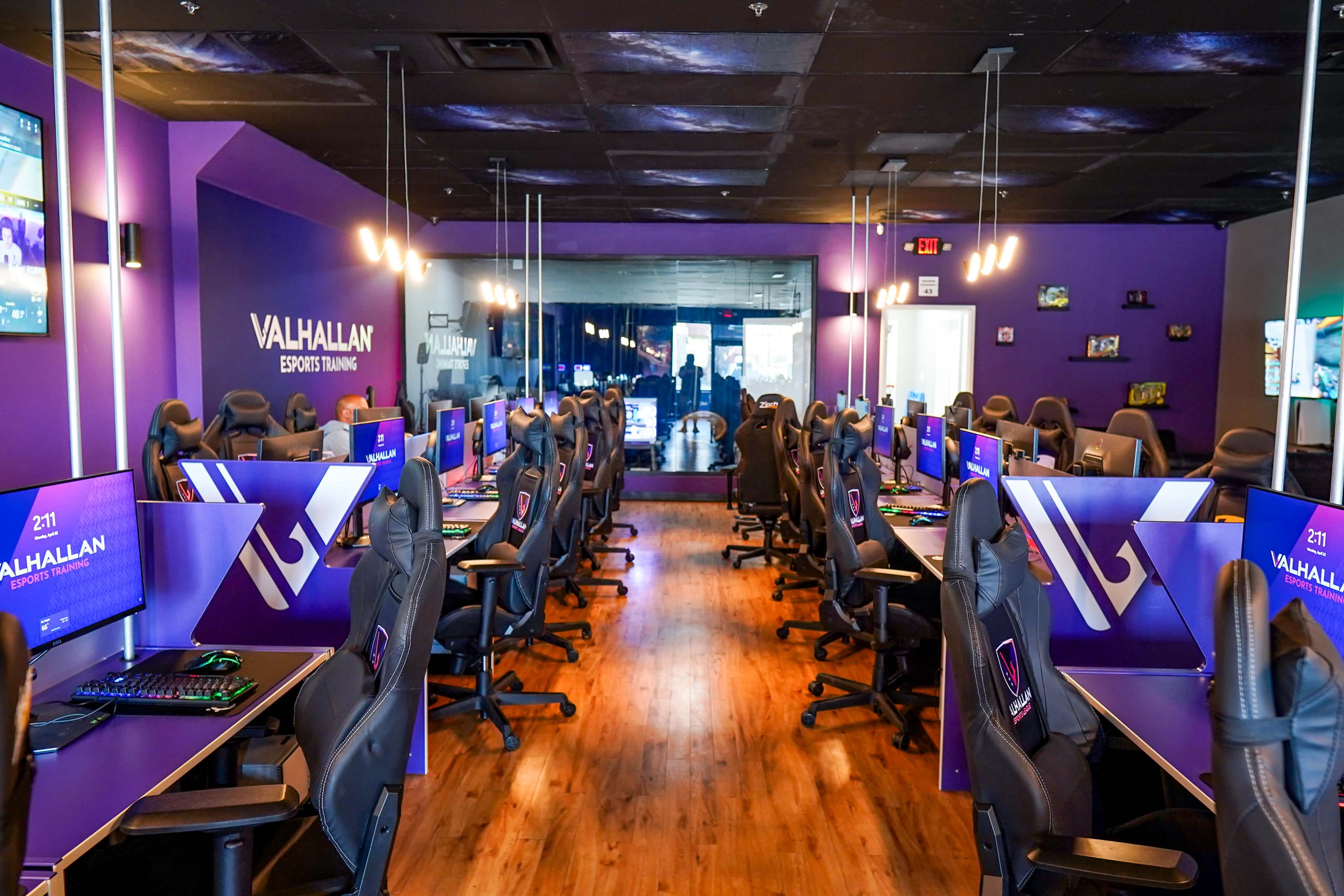 The image shows a gaming facility with rows of gaming chairs and computers set up in a purple-themed room, designed for esports training.