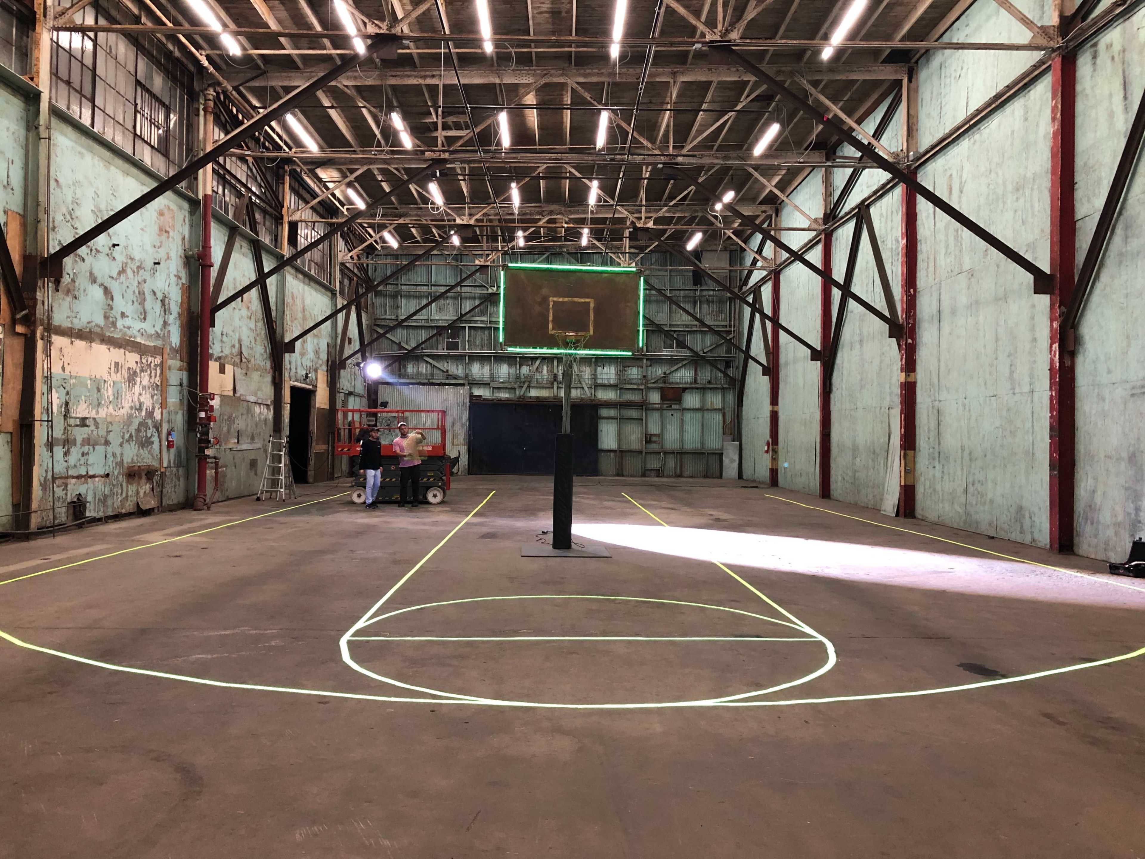 The image shows an empty indoor basketball court with highlighted court markings and a basketball hoop.