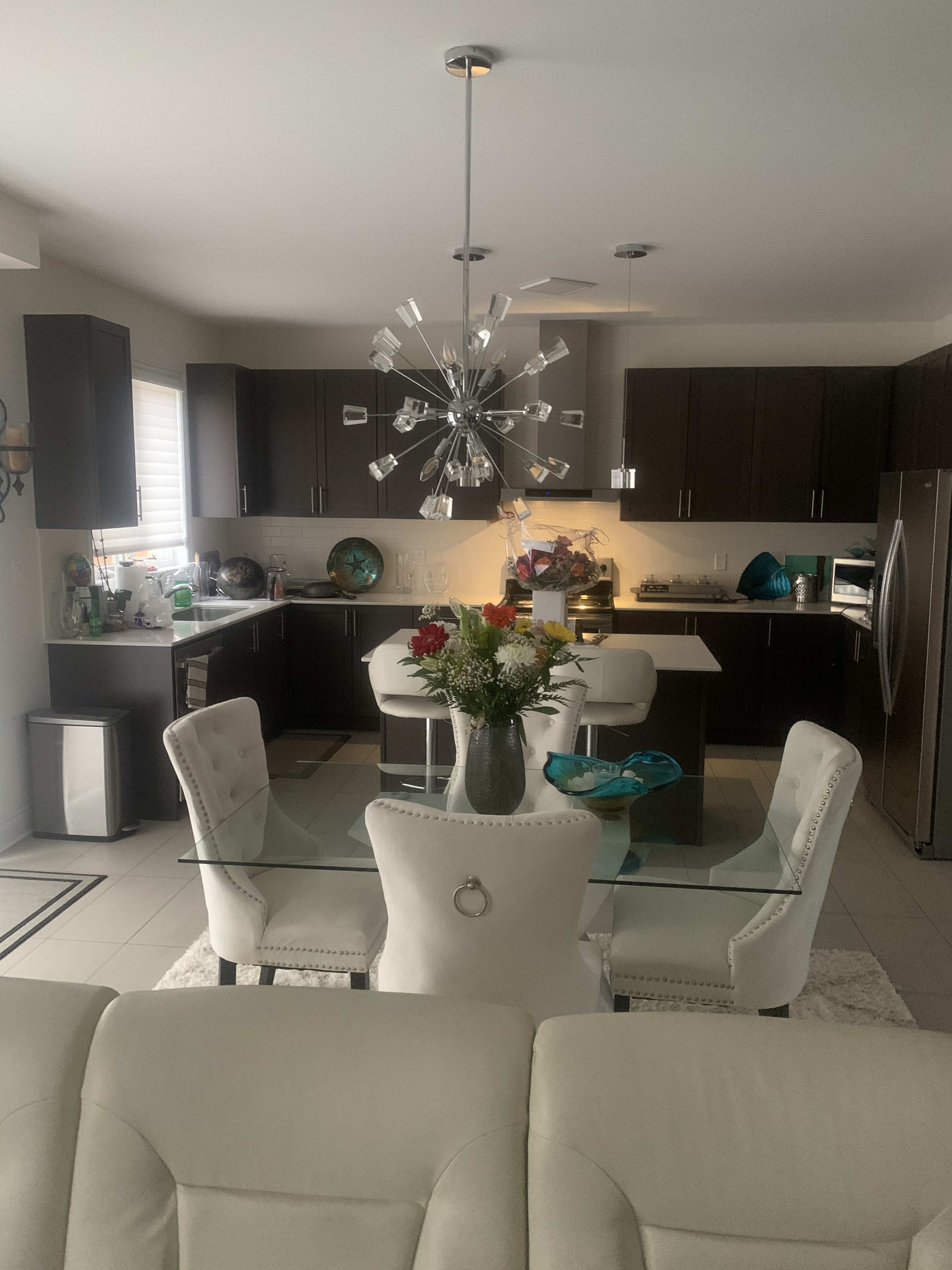 A modern kitchen features dark cabinetry, a glass dining table surrounded by white chairs, and a contemporary light fixture overhead.