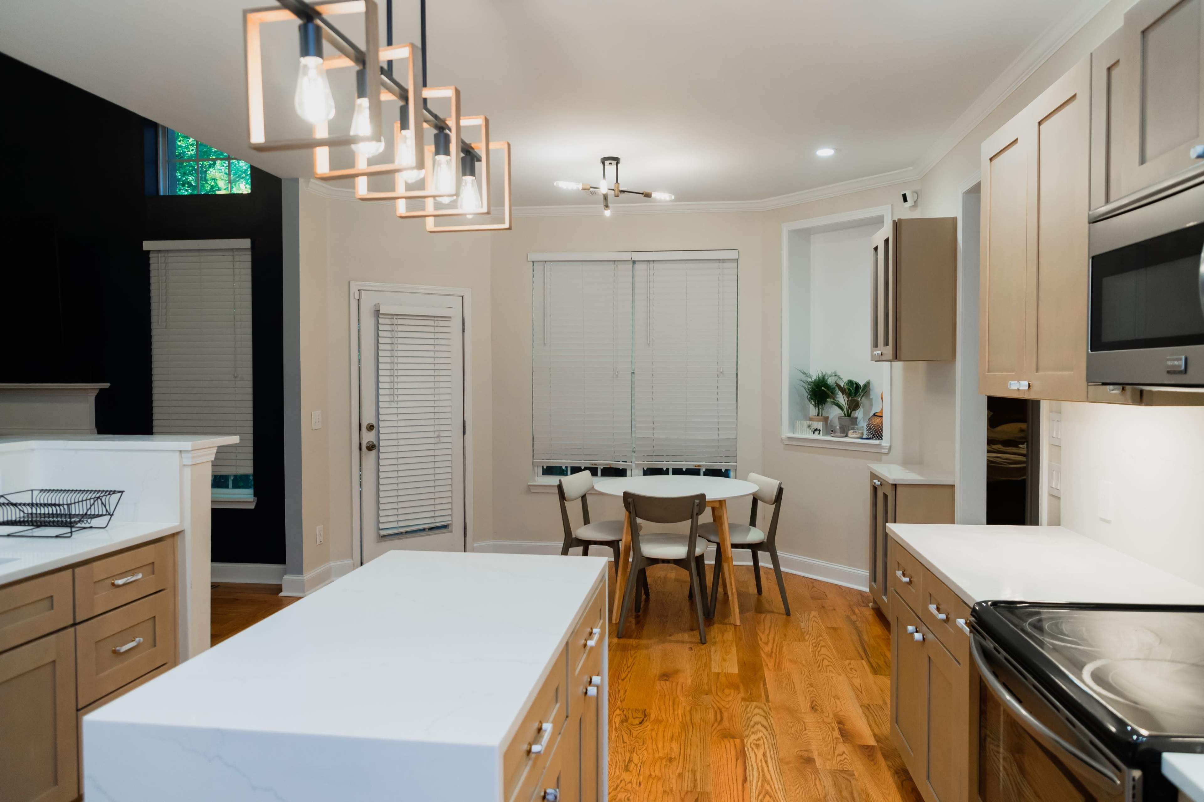 A modern kitchen with a central island, wooden cabinets, a dining table, and large windows allowing natural light to enter the space.