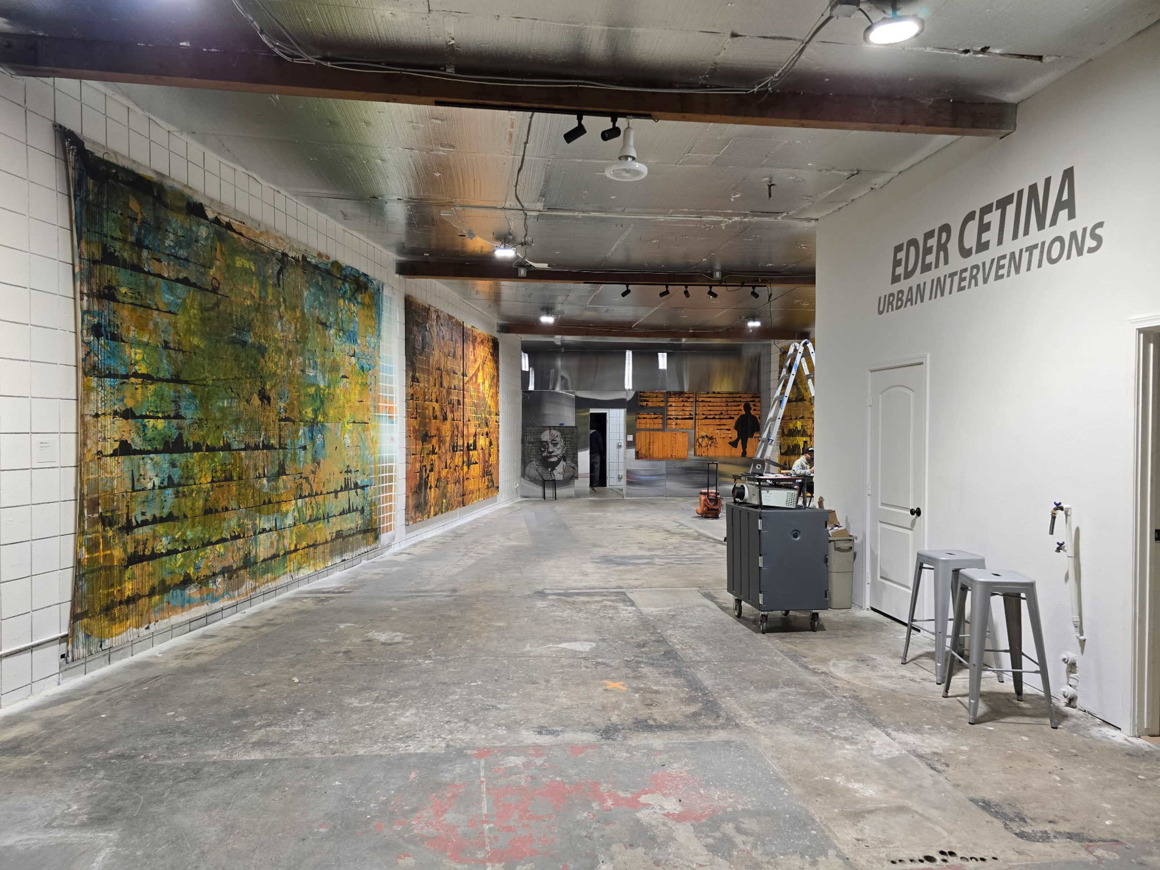 The image shows a spacious gallery with colorful abstract murals on the left wall and a gray artwork featuring text on the right, along with an installation area in the center.