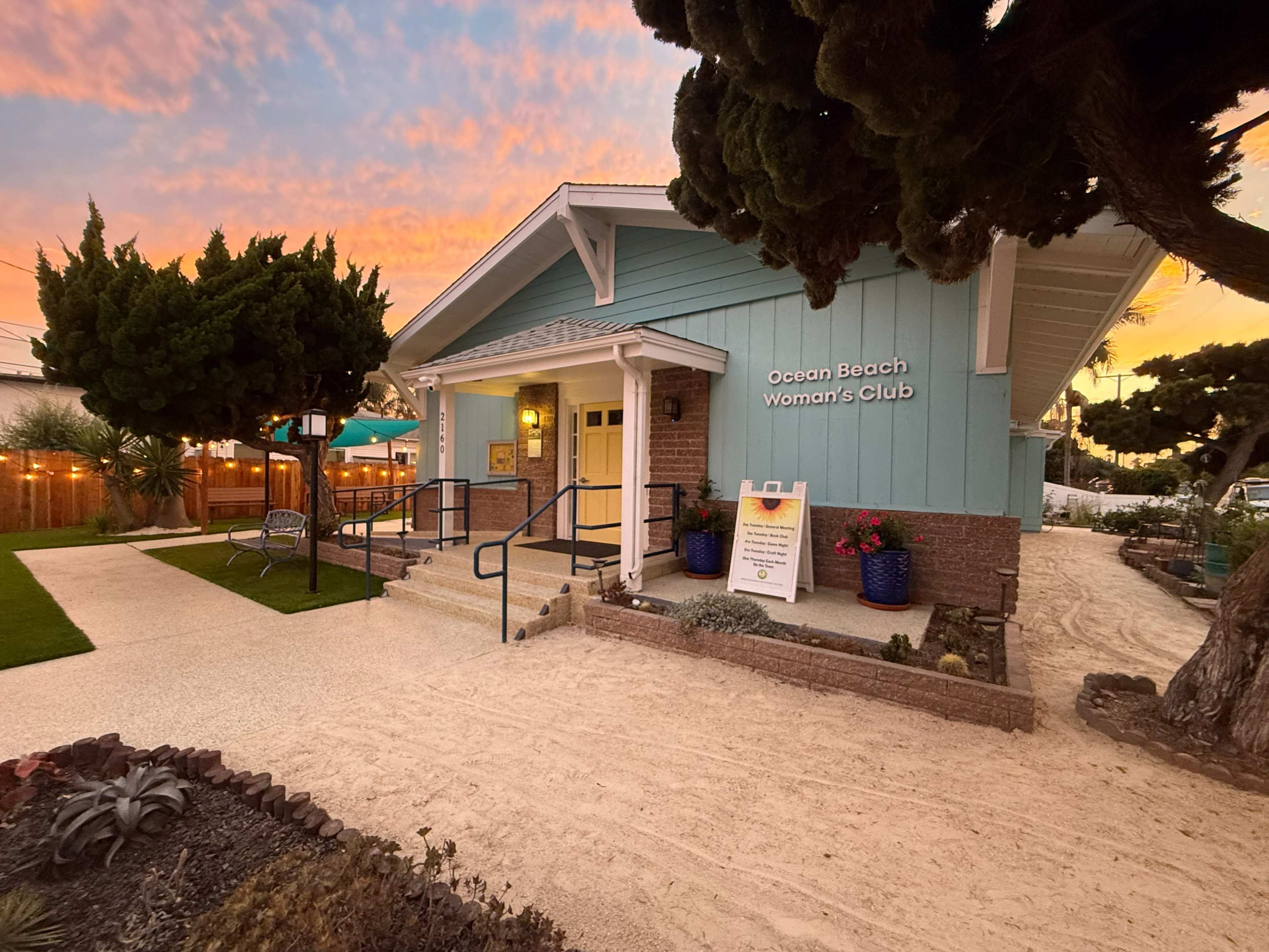 The image shows a light blue building with "Ocean Beach Woman's Club" signage, surrounded by landscaped greenery and a sandy pathway under a colorful sunset sky.