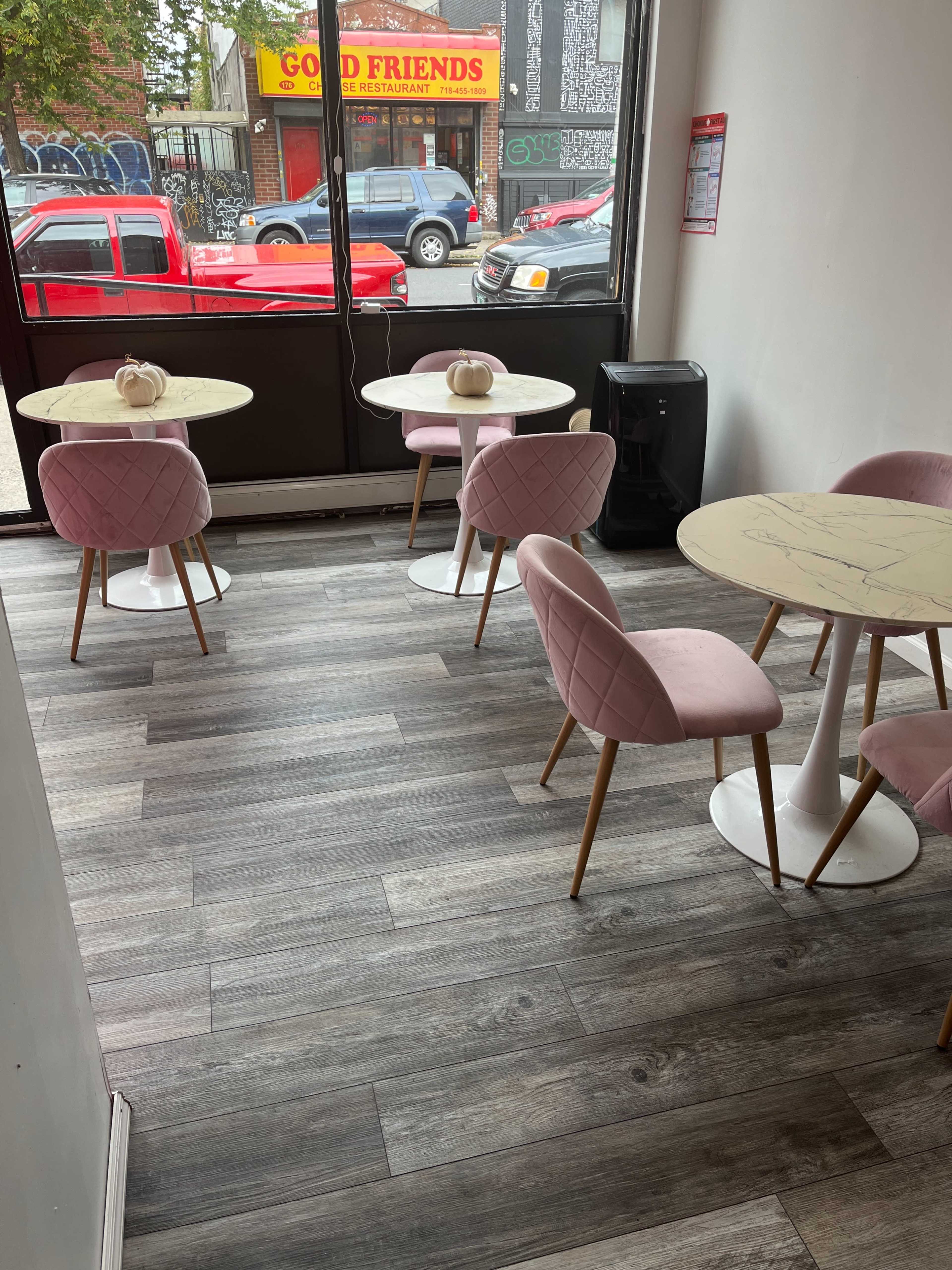 A modern café interior with pink chairs and round tables set on a wooden floor, featuring large windows that display a street view outside.