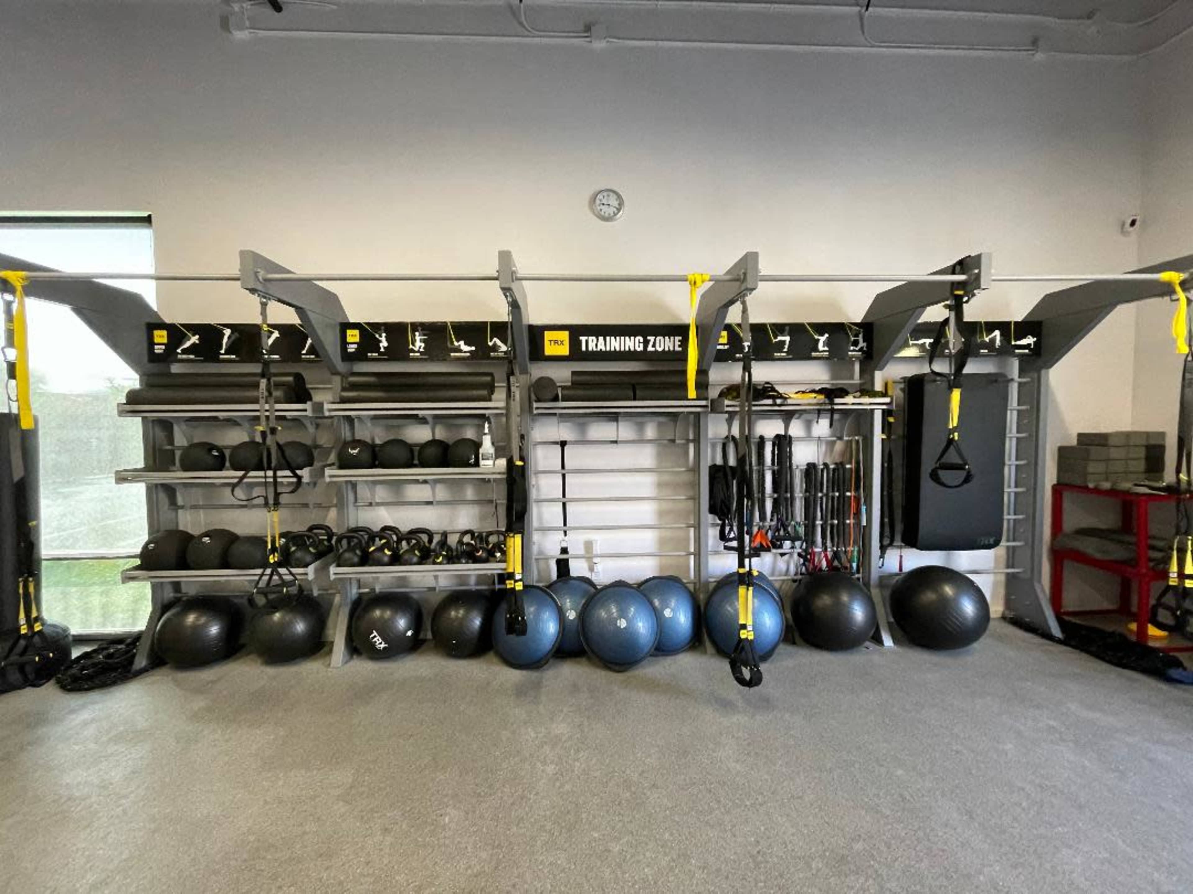 The image shows a well-organized gym area featuring various exercise equipment, including medicine balls, resistance bands, and suspension trainers arranged on shelves and the floor.