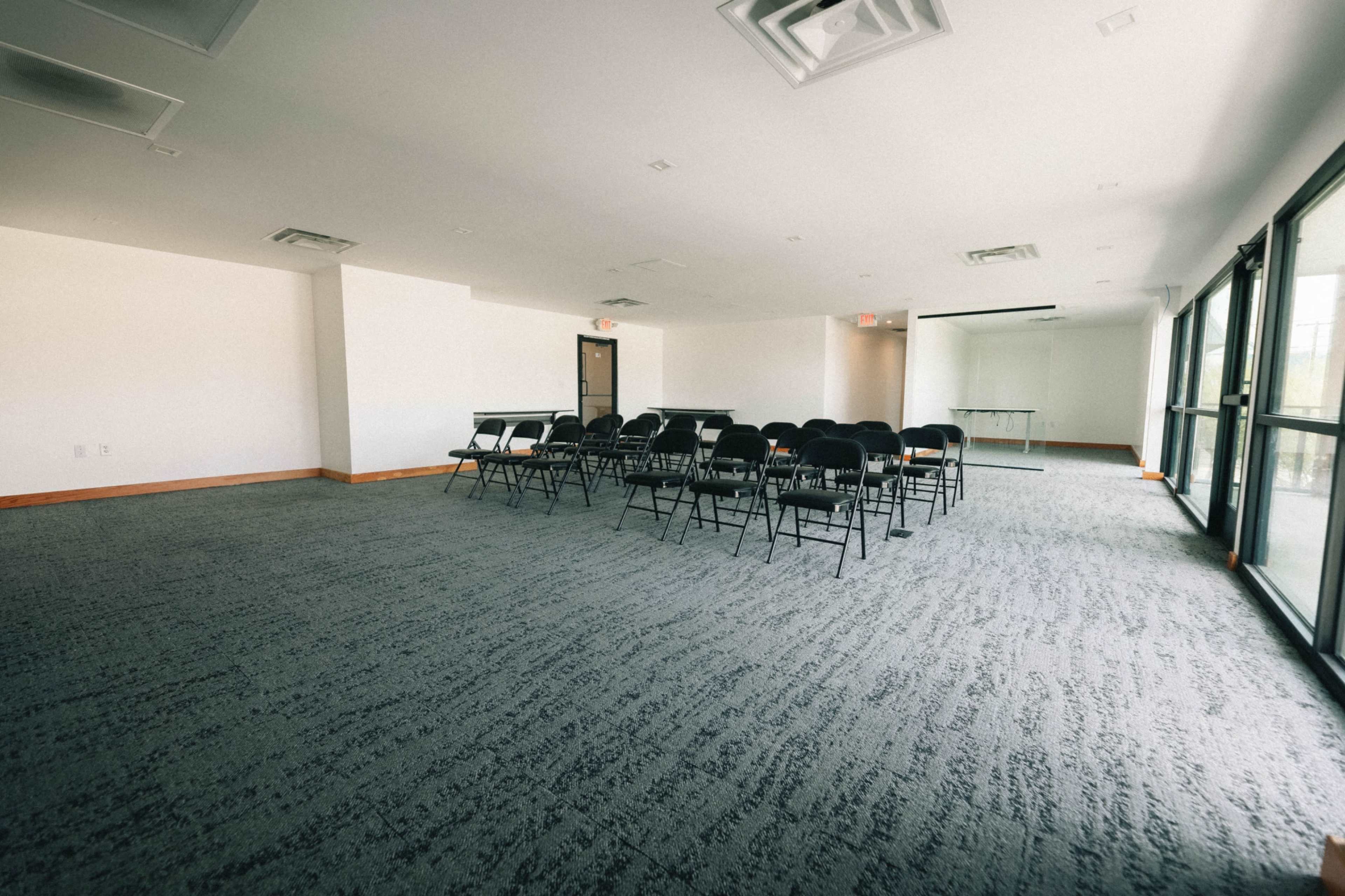 A large, empty meeting room features a set of black folding chairs arranged in rows on a carpeted floor, with windows on one side allowing natural light.