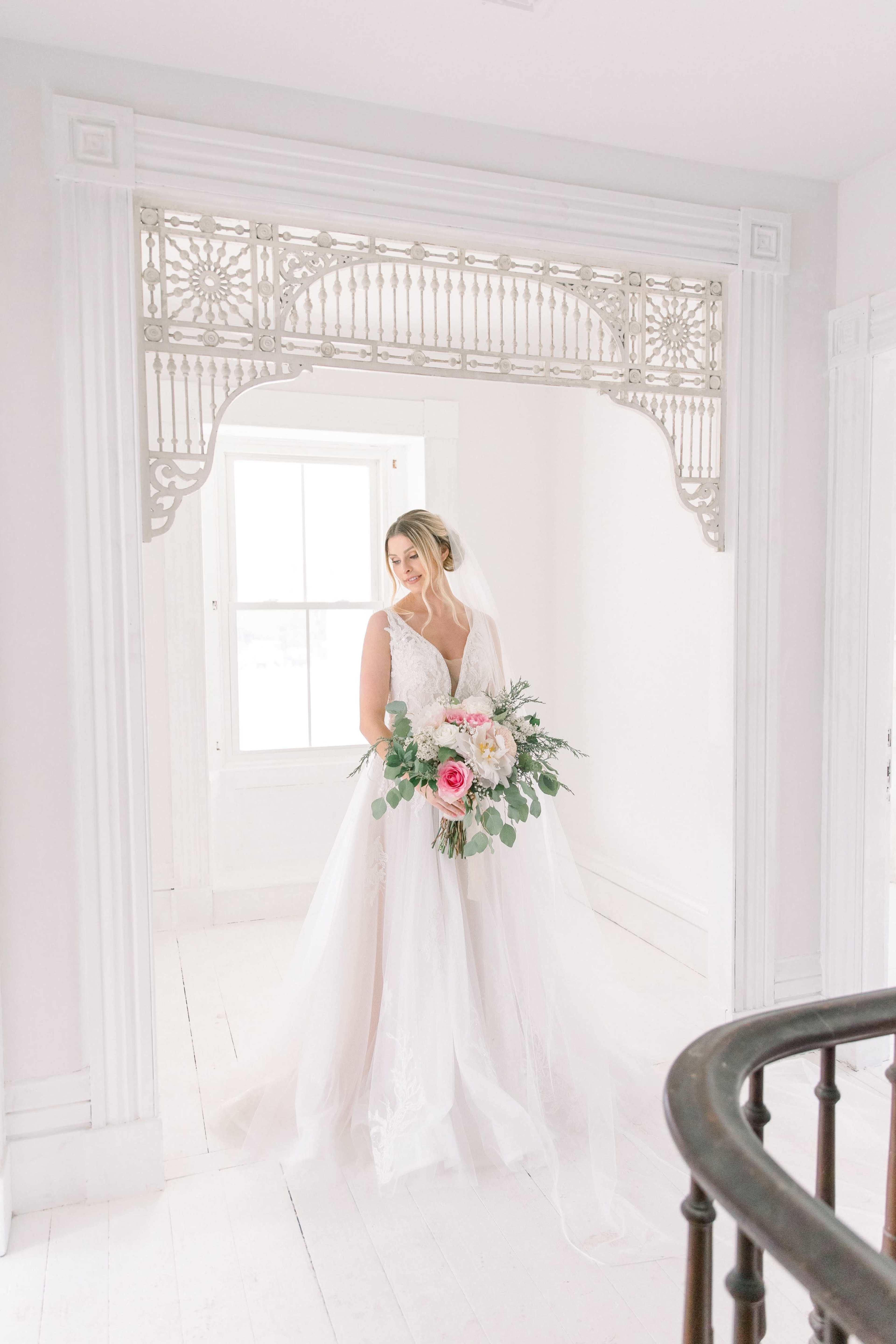 A bride holding a bouquet stands in a bright room adorned with ornate trim.