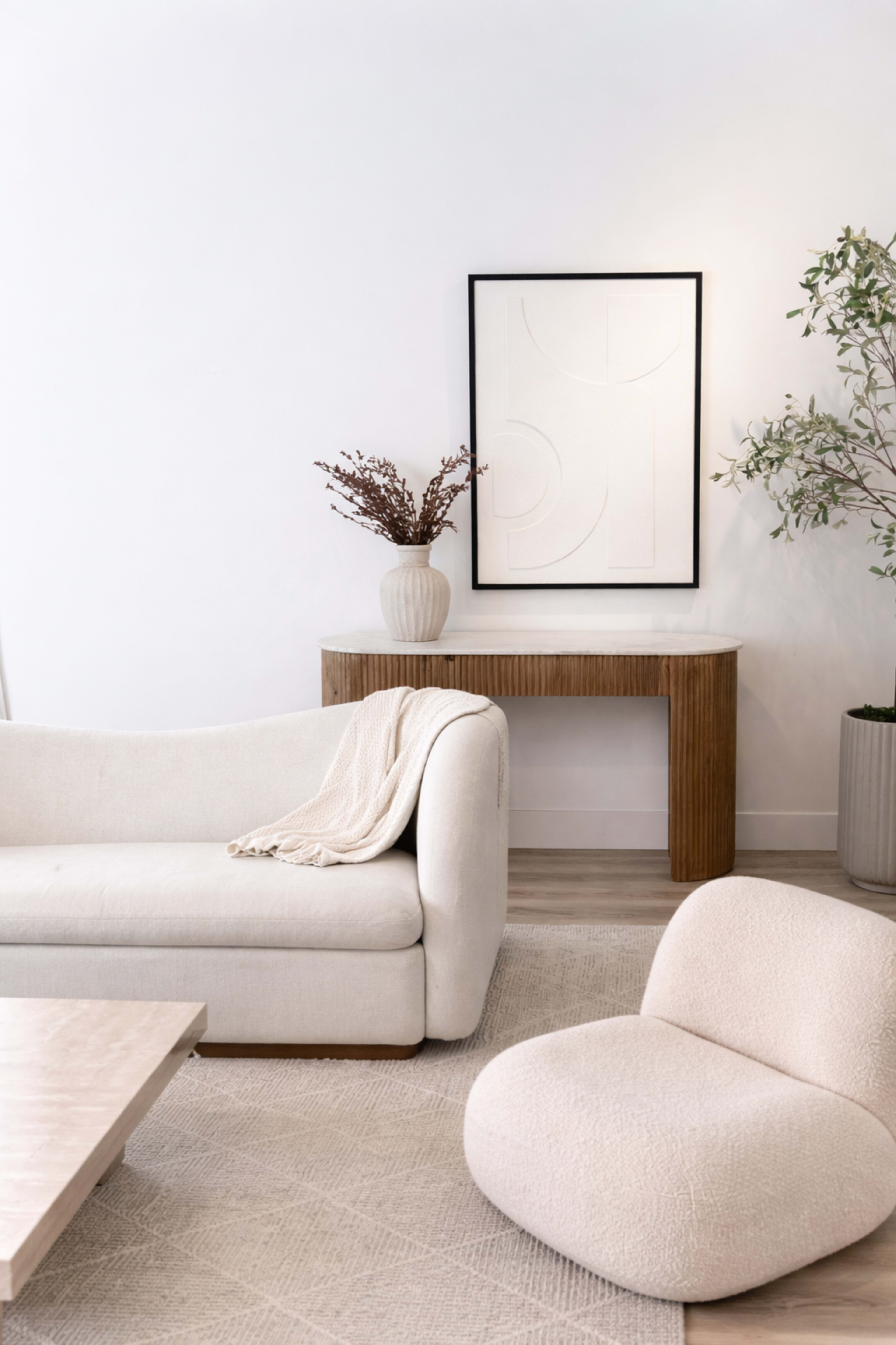 A light-colored living room features a textured sofa, a minimalist wooden console table with a decorative vase, and a plush armchair, all set against a white wall.