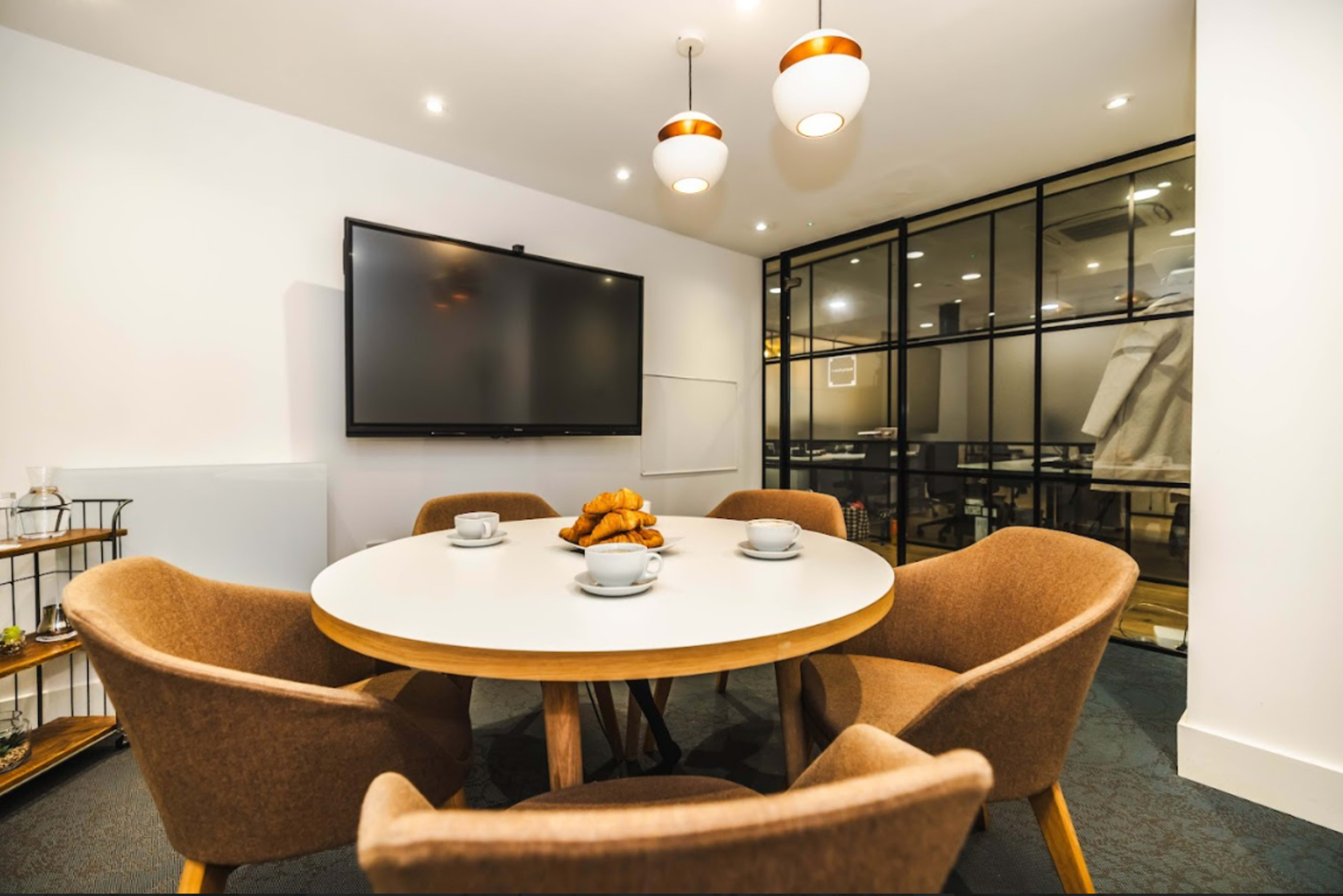 A round table with four upholstered chairs and a television mounted on the wall are arranged in a modern meeting room with a glass partition.