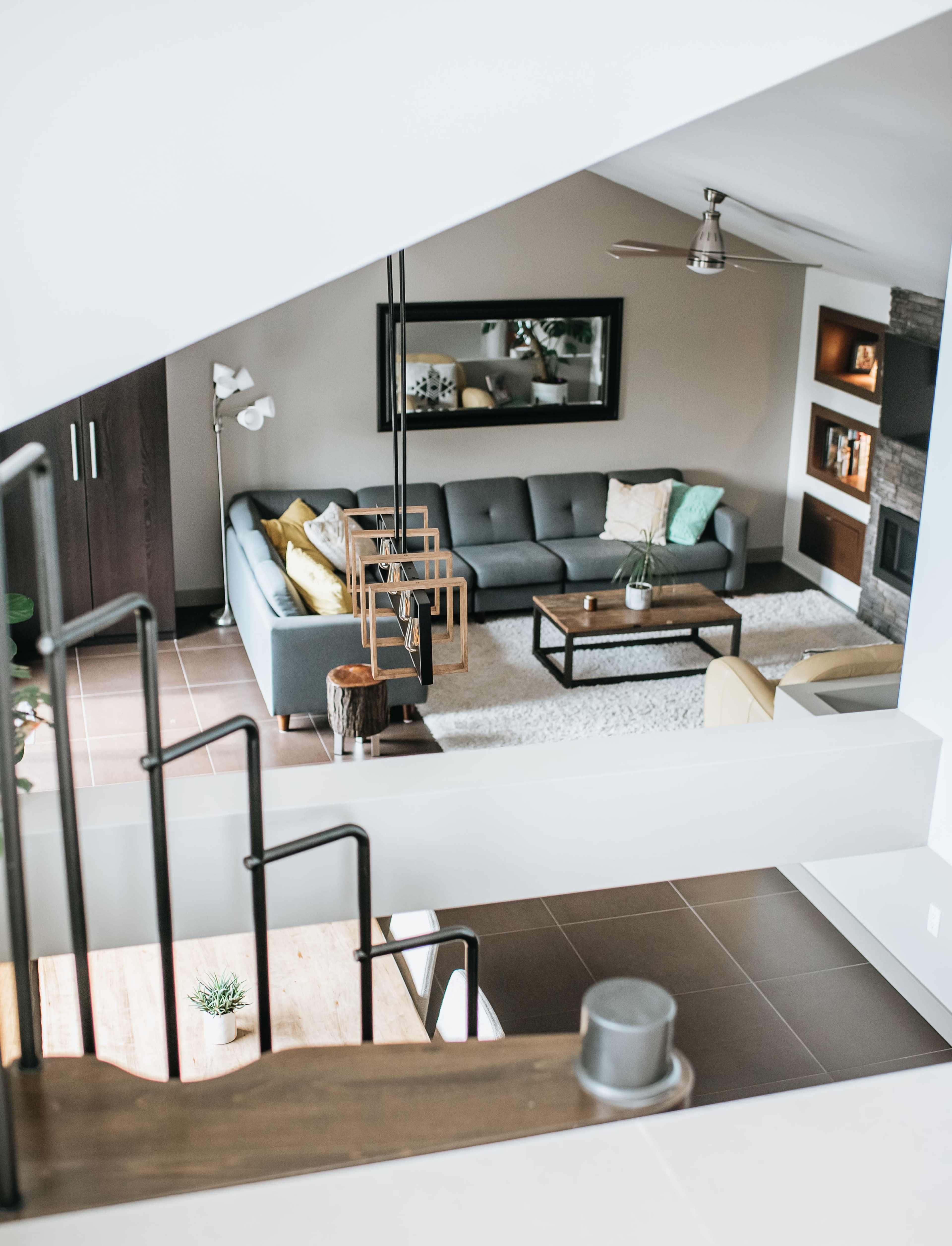 A modern living room features a gray sectional sofa, a coffee table, and a light rug, viewed from an upper landing with a staircase railing in the foreground.