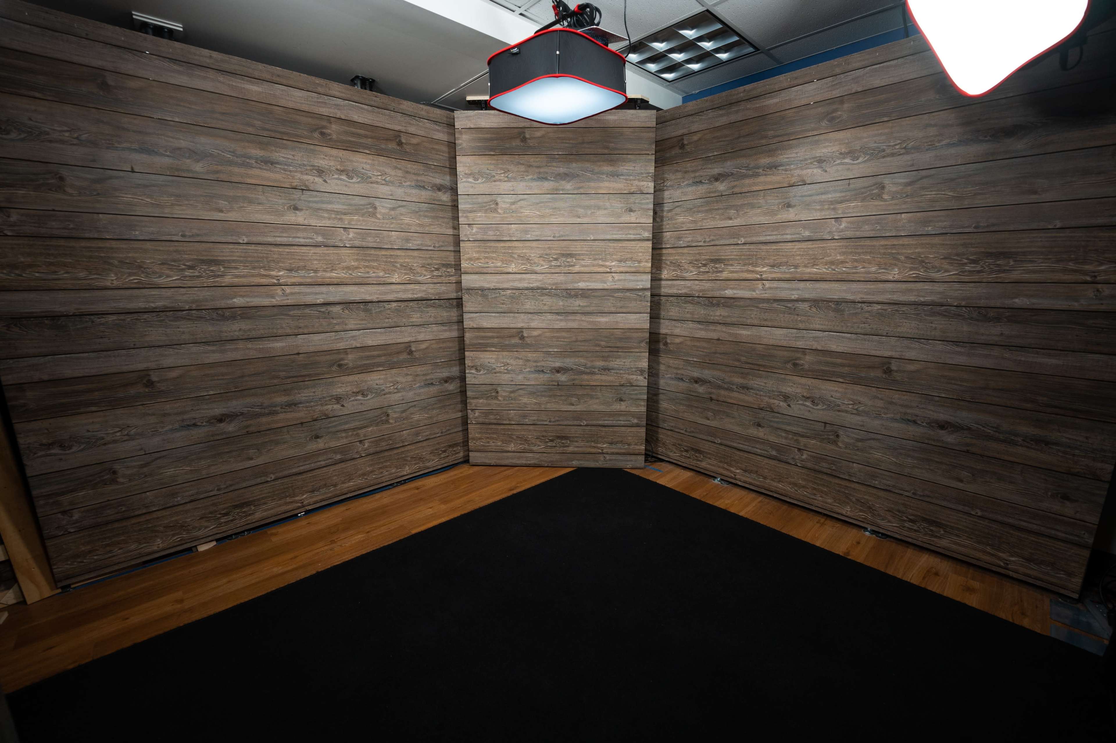 A studio setup with three wood-paneled backdrops positioned at angles, surrounded by a black carpeted floor.
