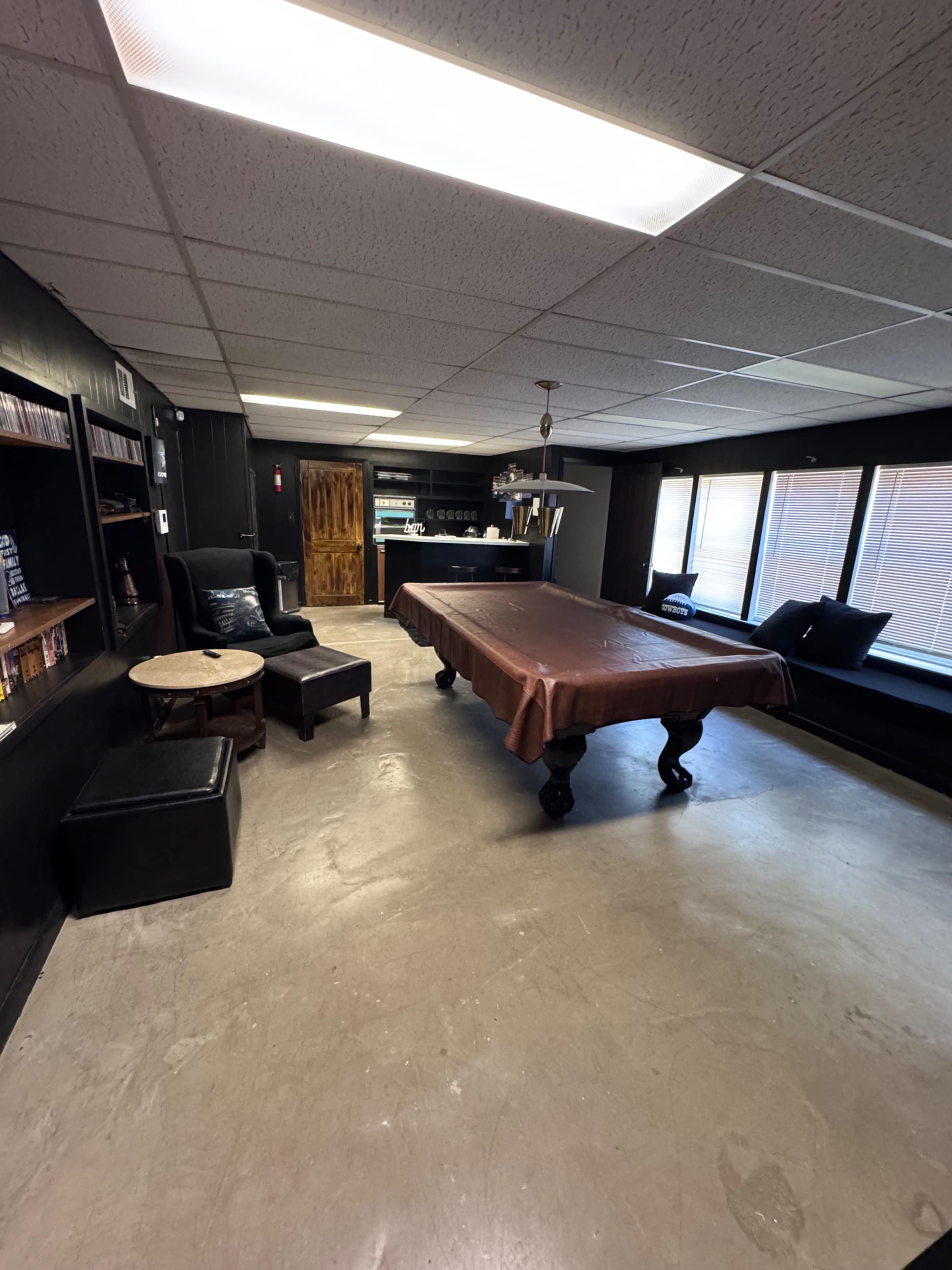 The image shows a dimly lit room featuring a billiard table in the center, surrounded by seating arrangements and shelves filled with books.