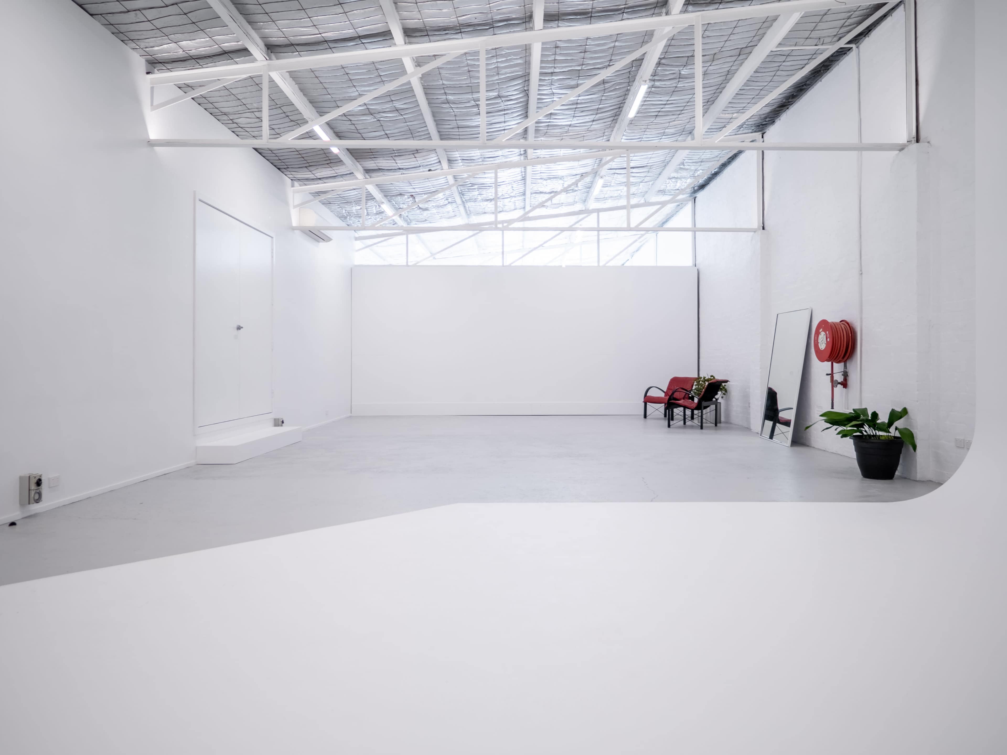 A spacious, minimalist studio features a white wall, a potted plant, a mirror, and a red fire extinguisher against the walls.